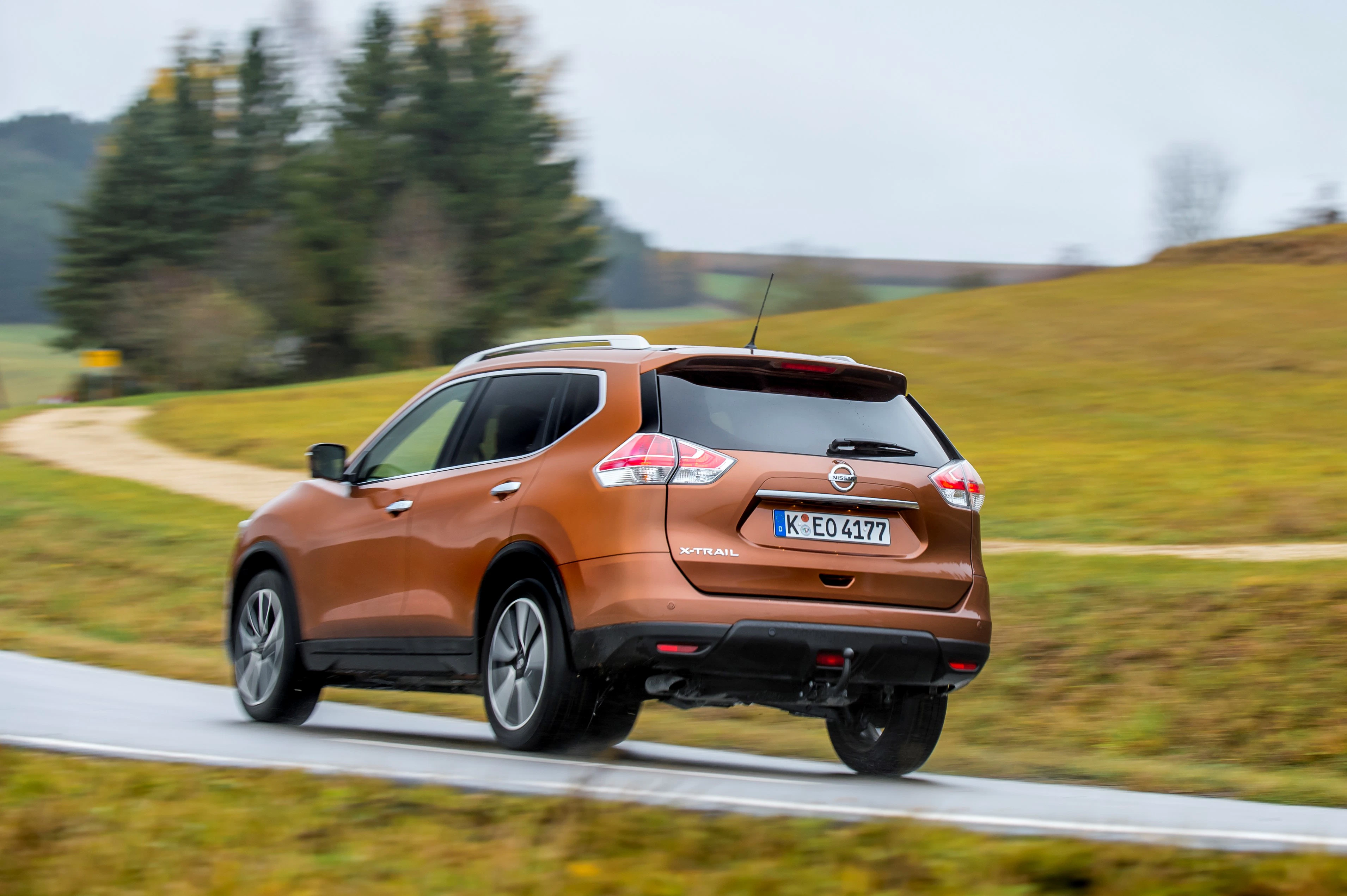 Brauner SUV fährt schnell auf einer ländlichen Straße, umgeben von grünen Feldern und Bäumen, bewölkter Himmel im Hintergrund.