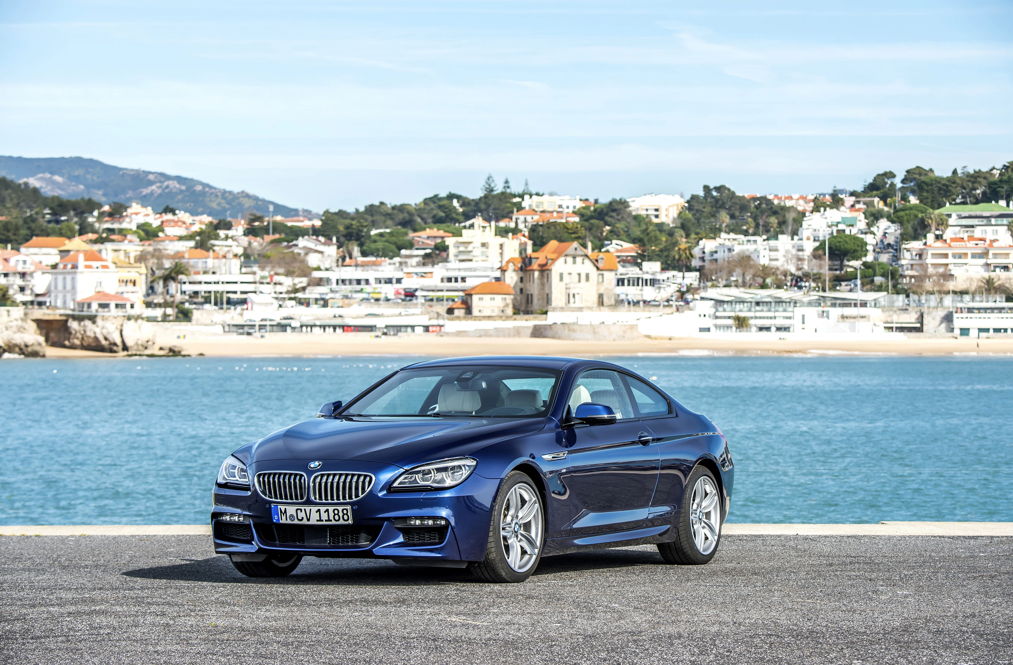 Ein blauer BMW steht auf einem Parkplatz vor einer Küstenstadt mit vielen weißen Häusern und einem ruhigen, blauen Meer im Hintergrund.