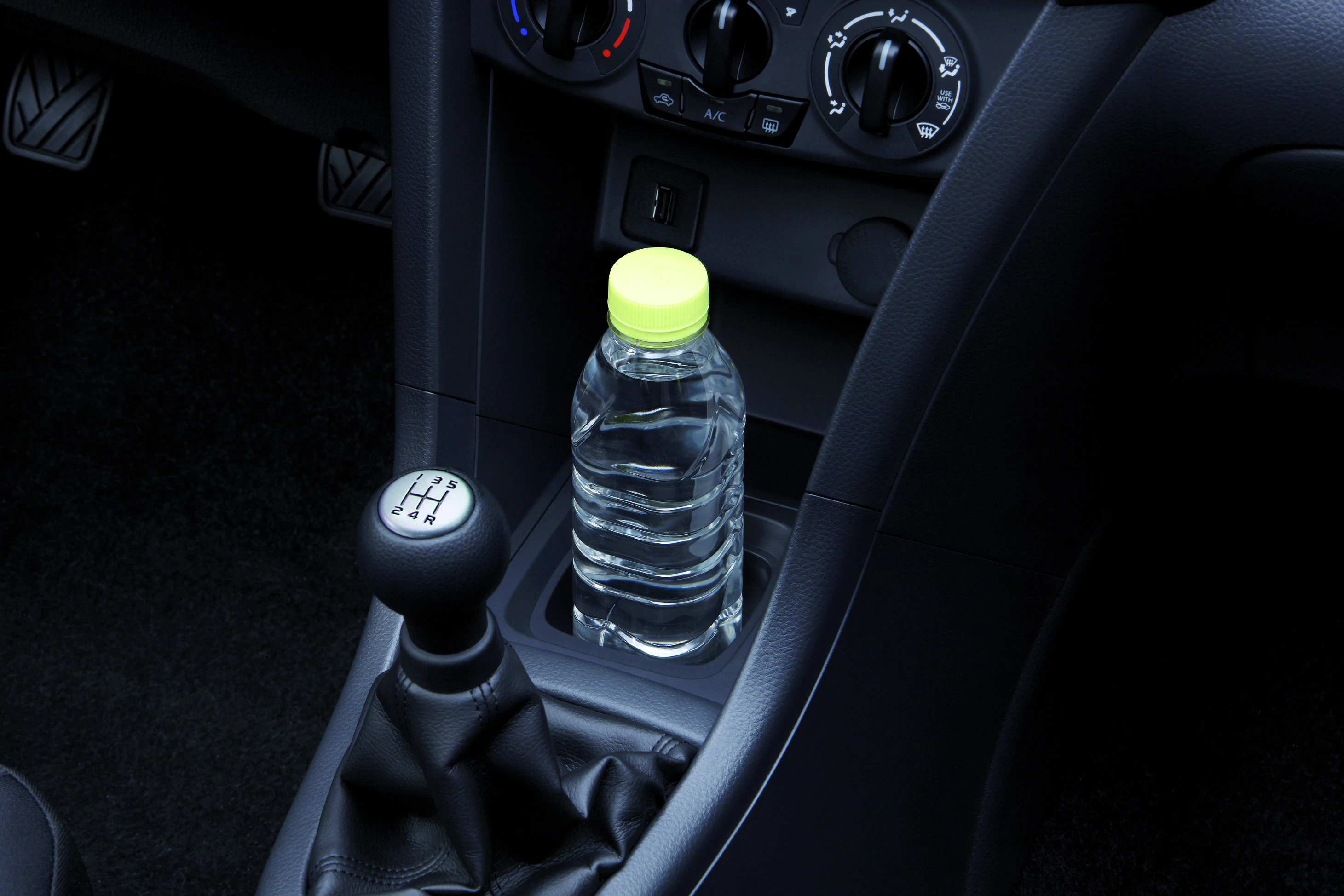 Nahaufnahme eines Autoschaltknüppels und einer Wasserflasche mit gelbem Verschluss im Getränkehalter der Mittelkonsole.