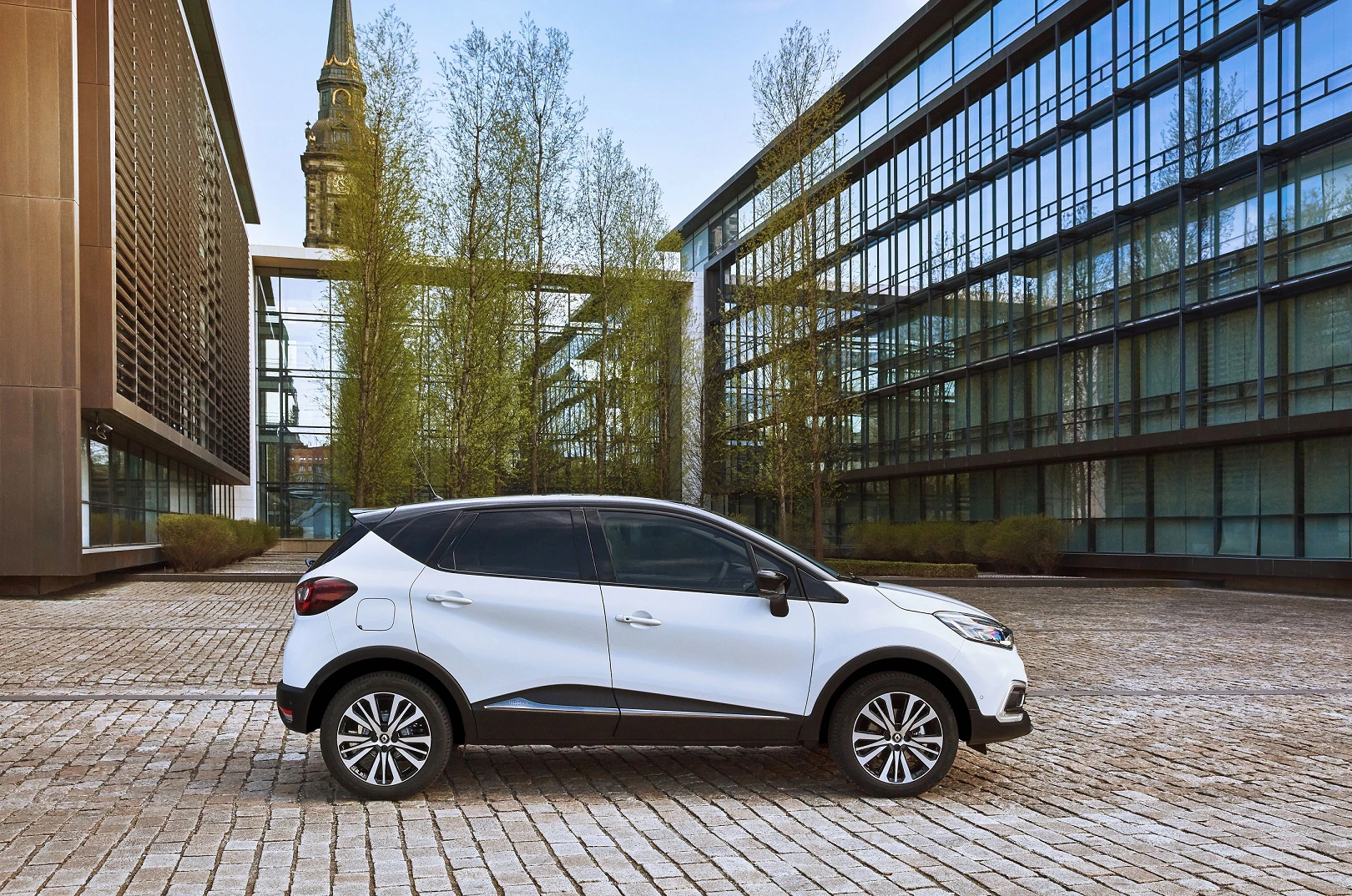 Weißes Auto auf gepflastertem Platz zwischen modernen Glasgebäuden und einem Kirchturm im Hintergrund.