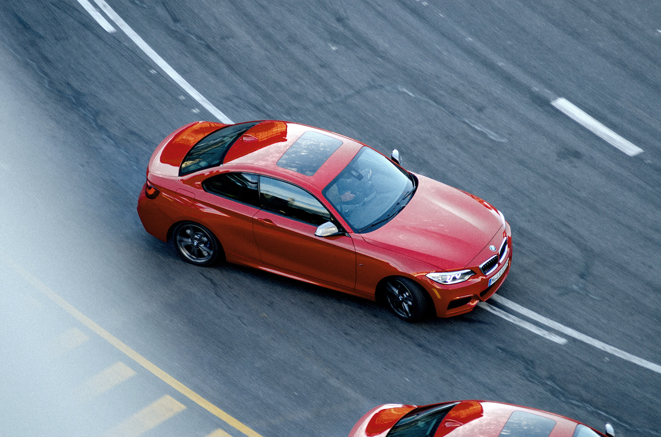 Roter Sportwagen in Kurvenfahrt auf der Straße, von oben fotografiert. Ein ähnliches Auto befindet sich im Vordergrund.