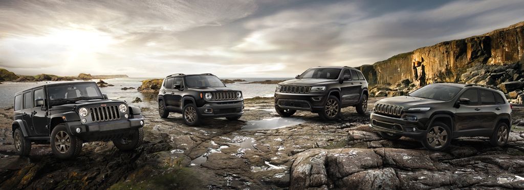 Vier schwarze Offroad-Fahrzeuge stehen auf felsiger Küste mit Meer und Himmel im Hintergrund. Ein Mensch auf einem Felsen hebt die Arme.