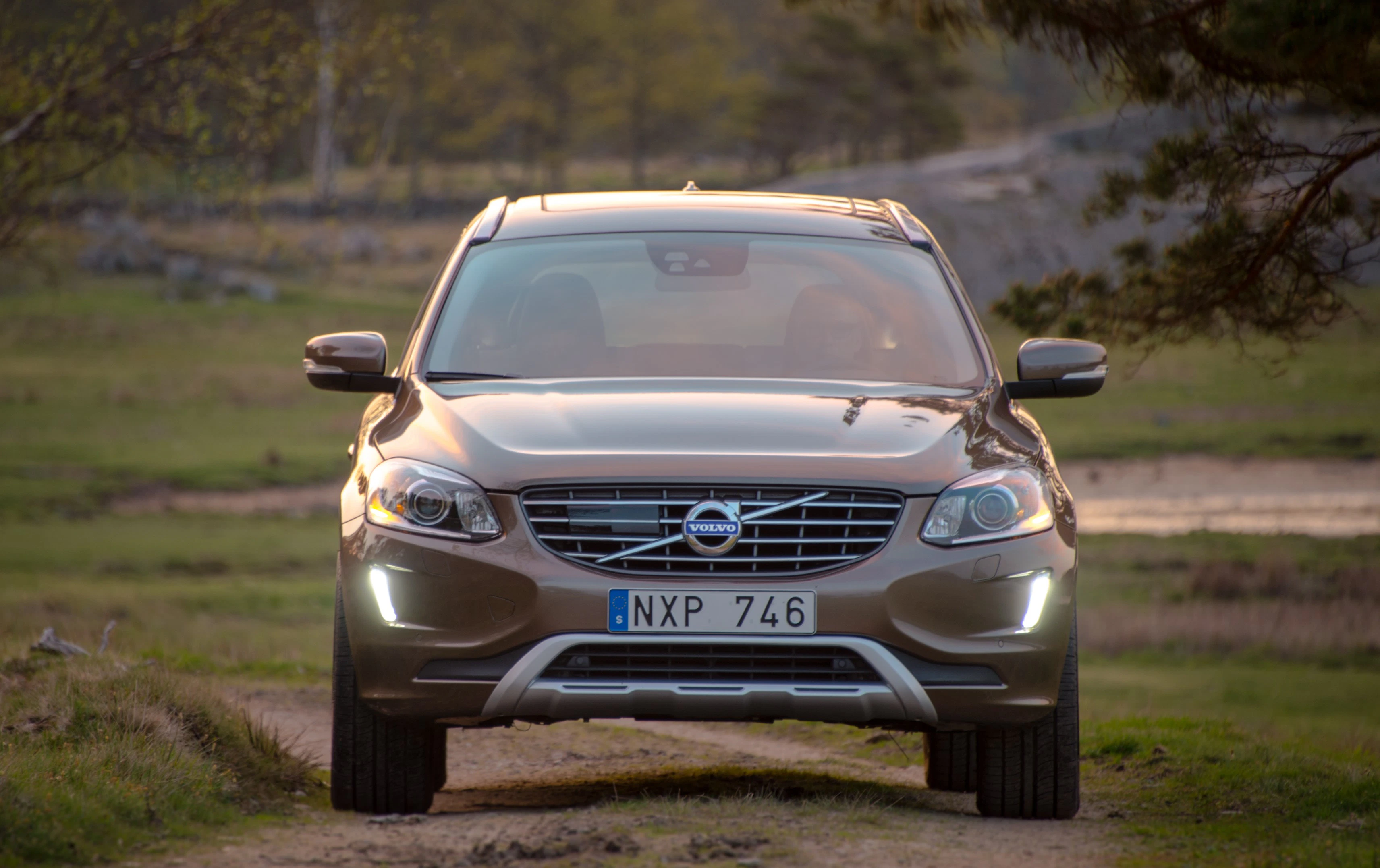 Ein brauner Volvo SUV fährt auf einem ländlichen Weg; Scheinwerfer sind eingeschaltet, Bäume und Wiesen im Hintergrund.