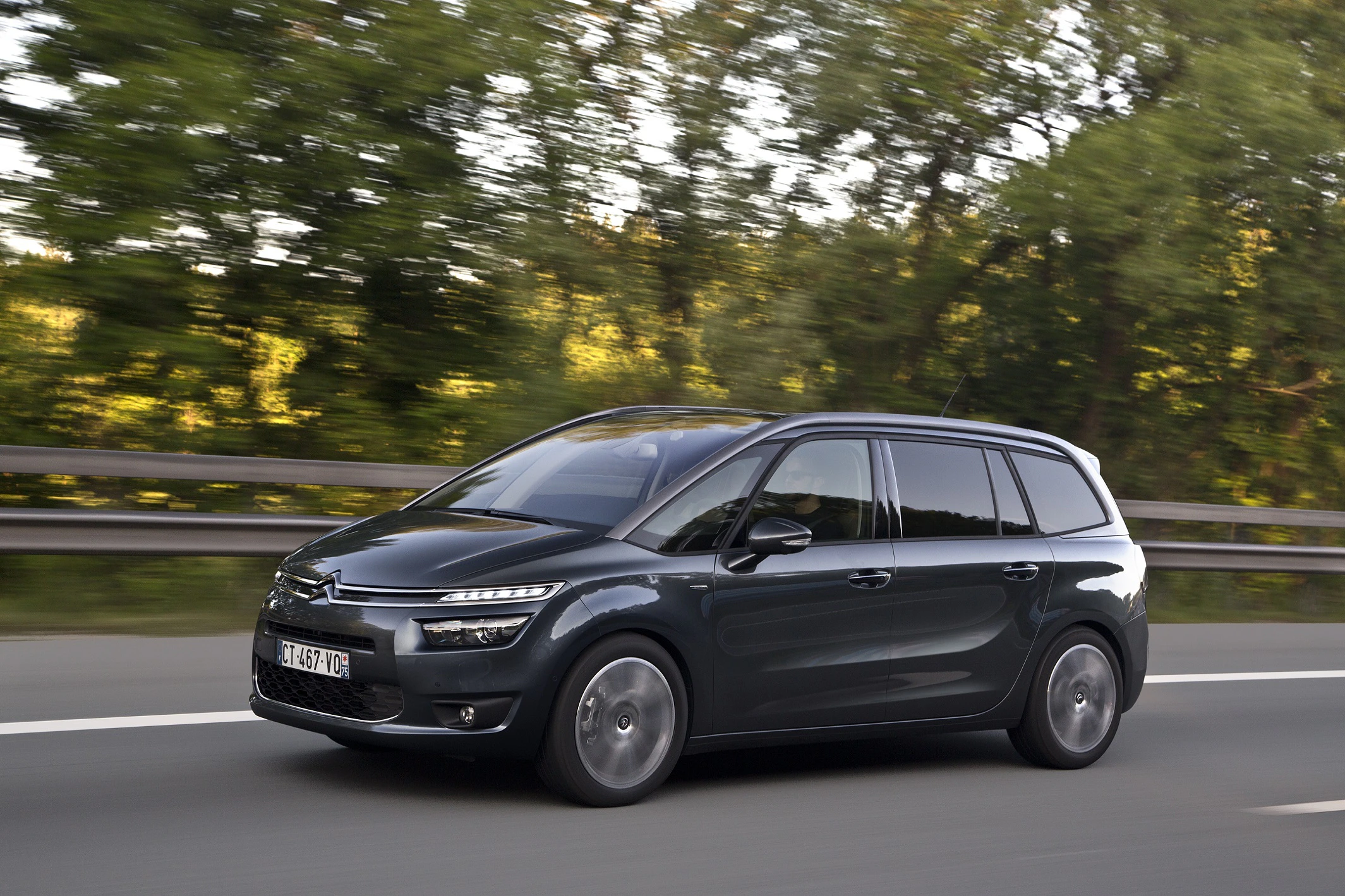 Ein dunkler Citroën C4 Grand Picasso fährt auf einer Straße, umgeben von Bäumen im Hintergrund.