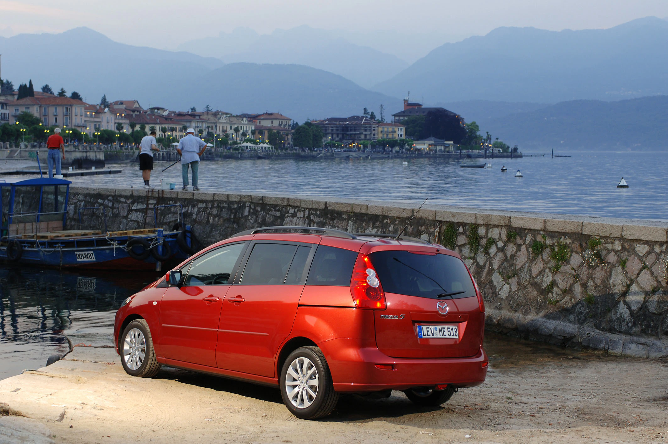 Rotes Auto parkt an einem Seeufer bei Dämmerung, im Hintergrund sind Berge und eine beleuchtete Stadt zu sehen.