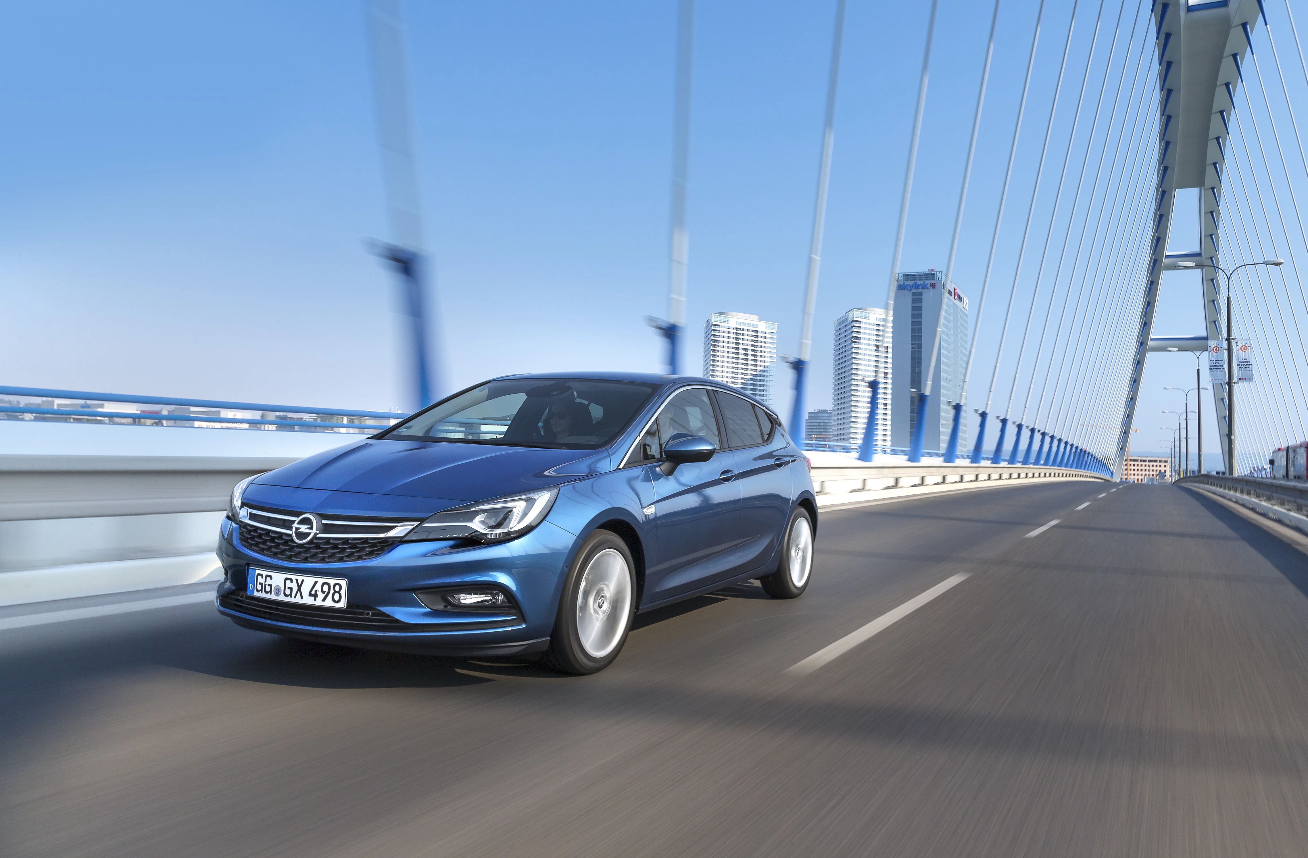 Blauer Opel Astra fährt über eine moderne Brücke, im Hintergrund sind Hochhäuser vor blauem Himmel zu sehen.