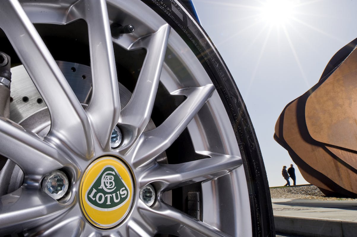 Nahaufnahme eines Lotus-Autoreifens in silberner Felge, im Hintergrund eine moderne Skulptur und zwei Personen, Sonnenlicht strahlt stark.