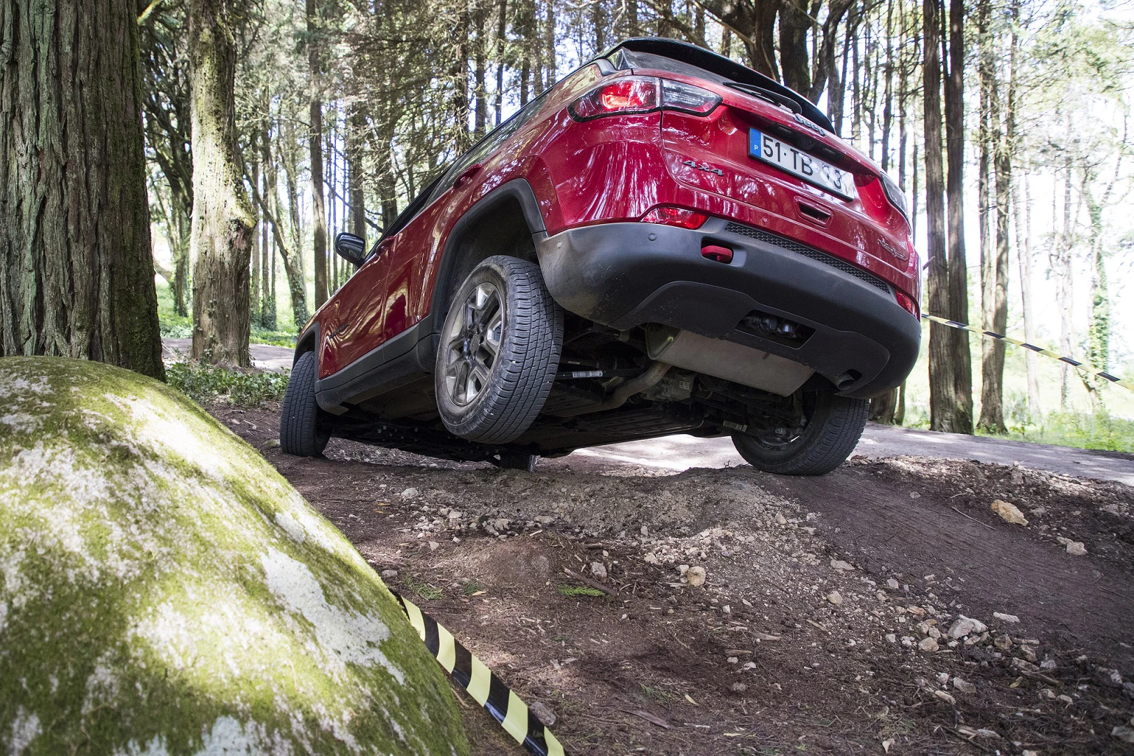 Roter SUV bewältigt schwieriges Gelände im Wald, ein Rad ist in der Luft. Boden uneben, Bäume im Hintergrund.