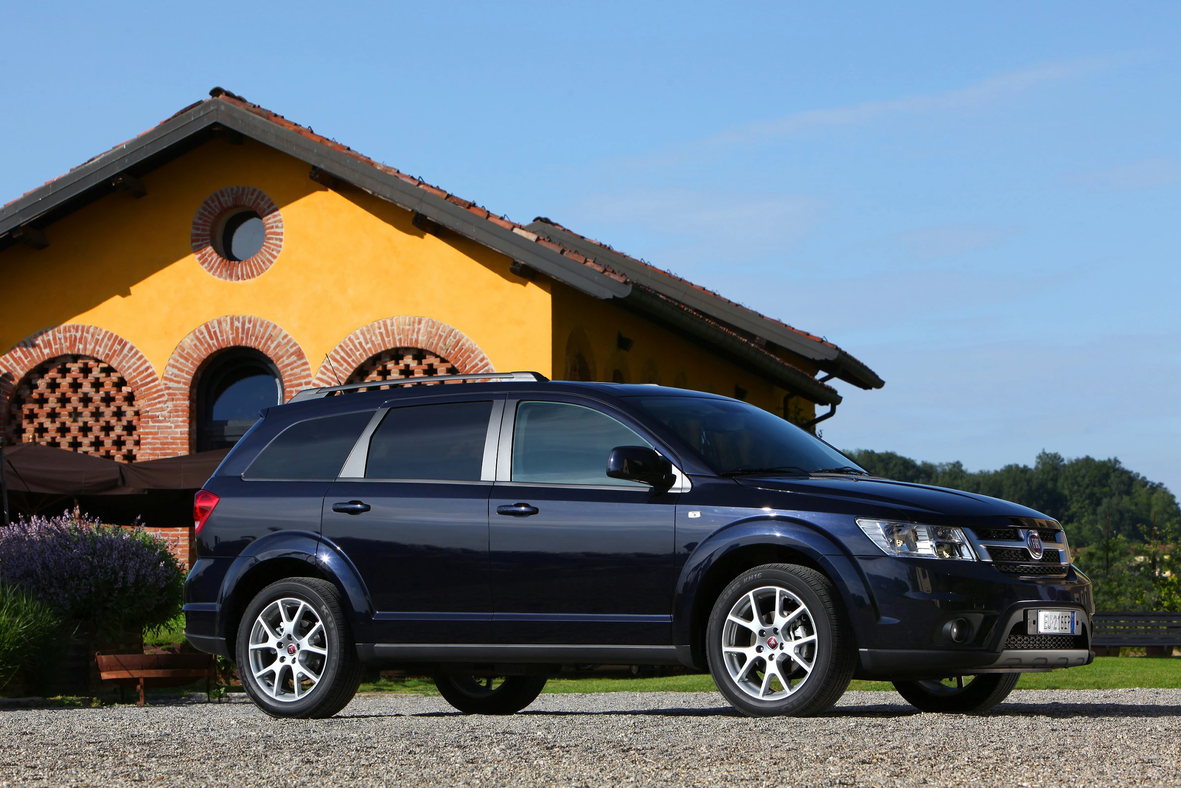 Dunkelblauer SUV parkt vor einem gelben Haus mit roten Ziegeln und Bogenfenstern, umgeben von grüner Natur unter blauem Himmel.