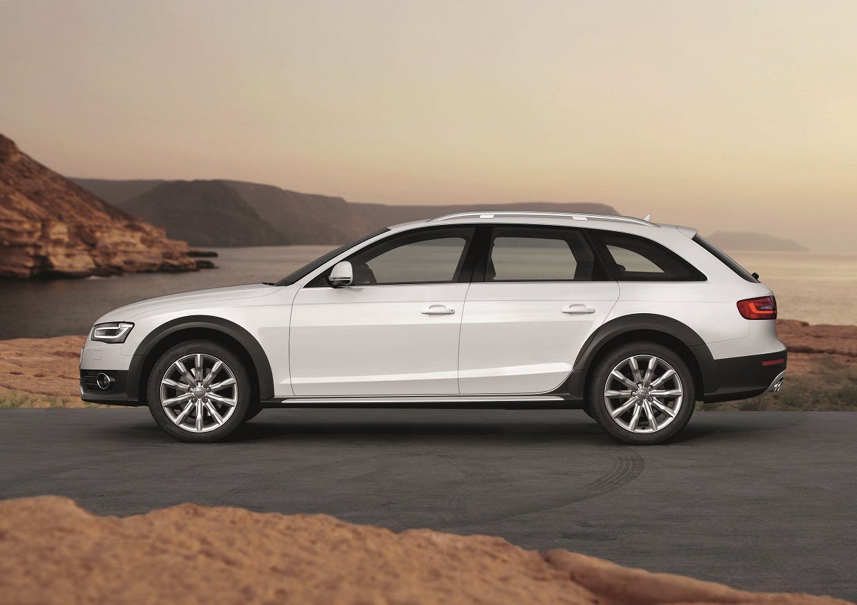 Weißer Kombi auf einer Küstenstraße bei Sonnenuntergang, umgeben von Felsen und Meer im Hintergrund.