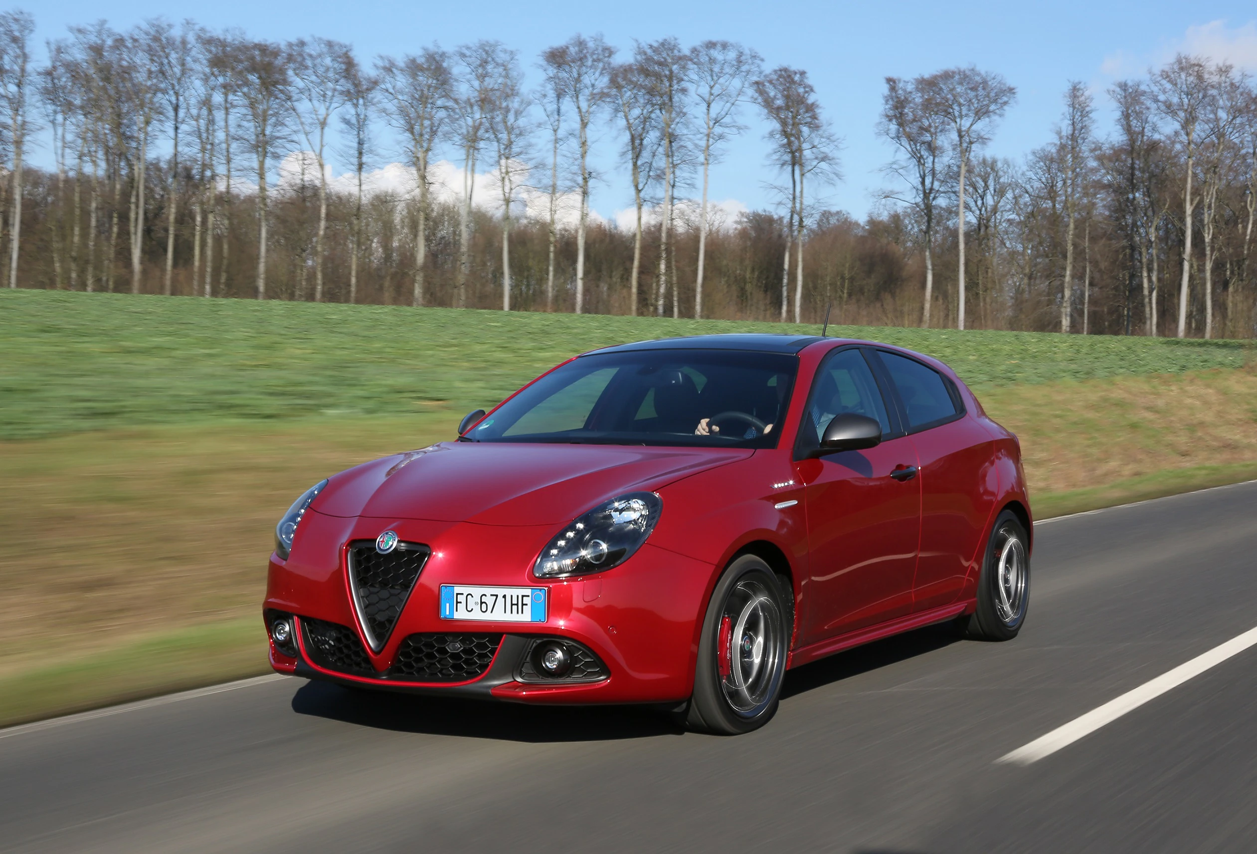 Roter Alfa Romeo Giulietta fährt auf einer Landstraße, umgeben von grünen Feldern und kahlen Bäumen an einem klaren Tag.
