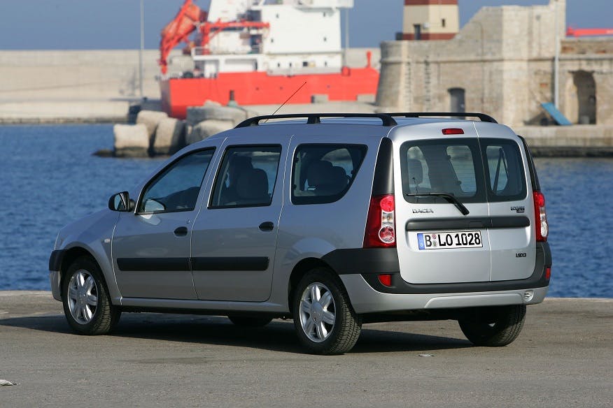 Silberner Dacia-Kombi parkt am Hafen vor einem roten Frachtschiff und Wasser im Hintergrund.