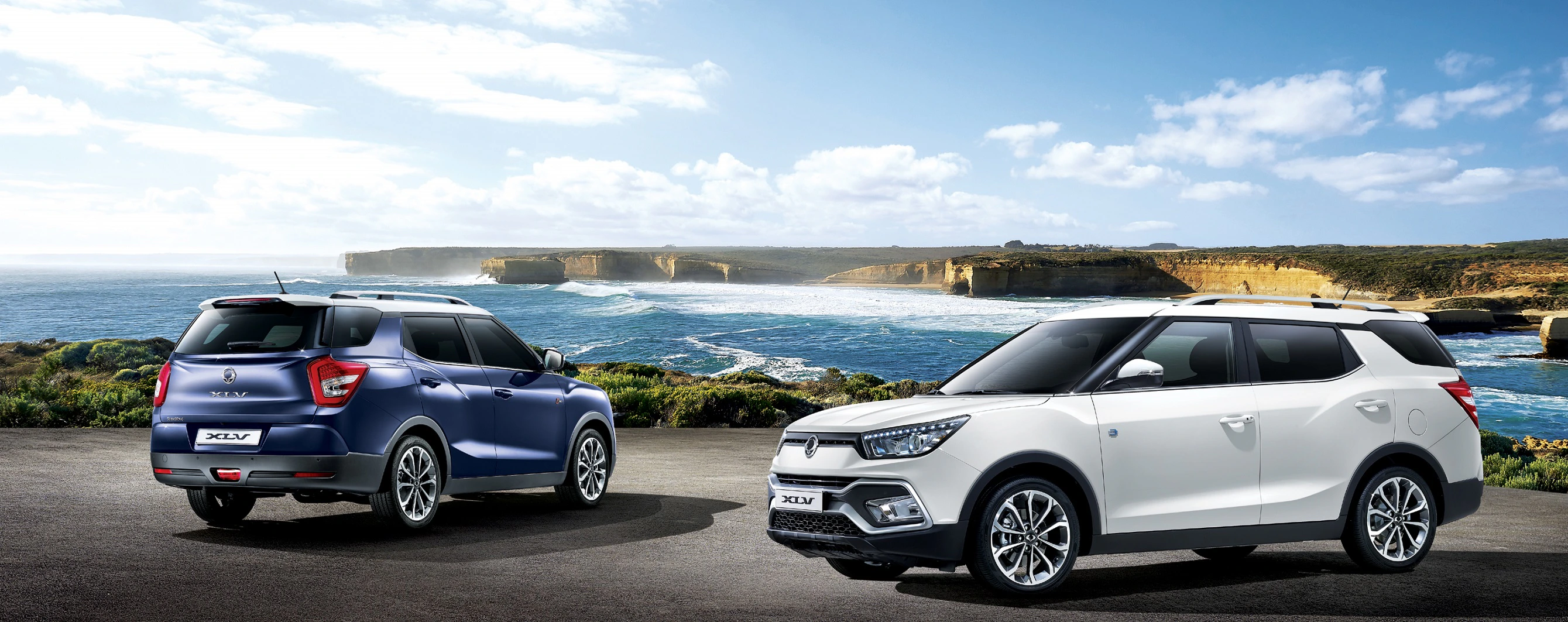 Zwei SUVs parken vor einer Küstenlandschaft mit blauem Himmel und Meer im Hintergrund, einer in Blau, der andere in Weiß.