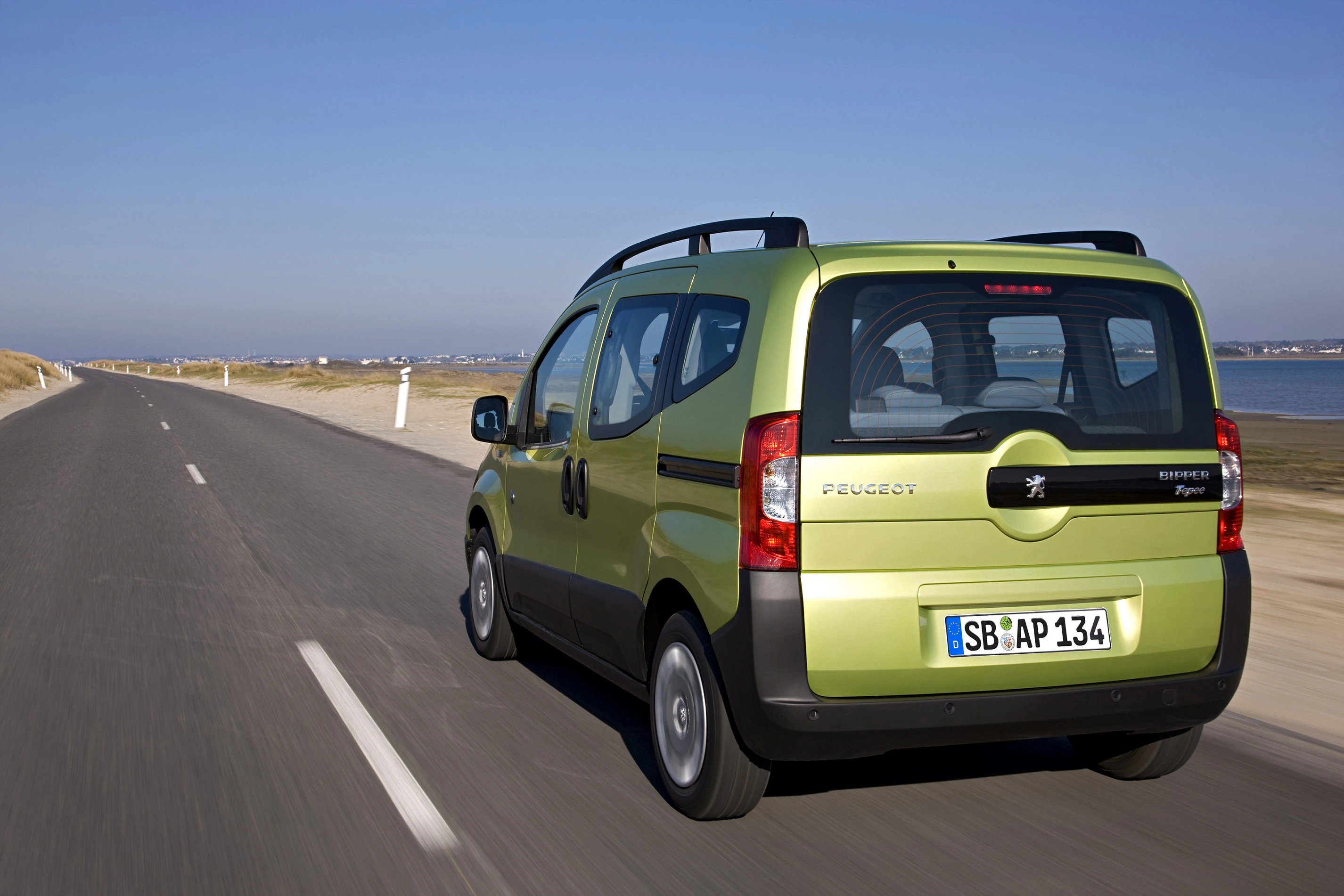 Grüner Peugeot-Kleintransporter fährt auf Landstraße entlang eines Gewässers bei klarem Himmel.