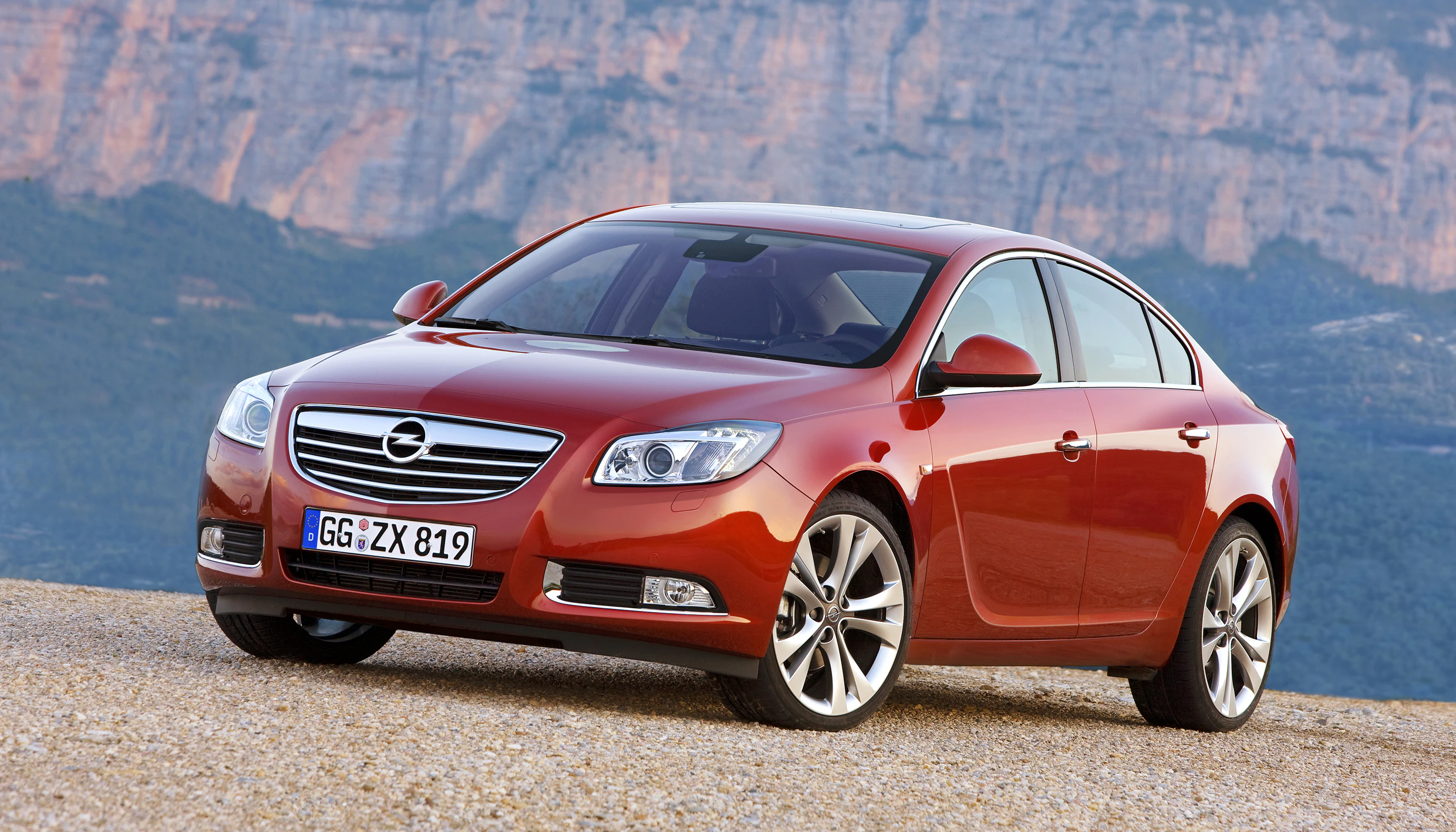 Roter Opel Insignia Limousine vor einer Berglandschaft, geparkt auf Kiesweg. Vorderansicht mit deutlichem Markenlogo und Nummernschild.