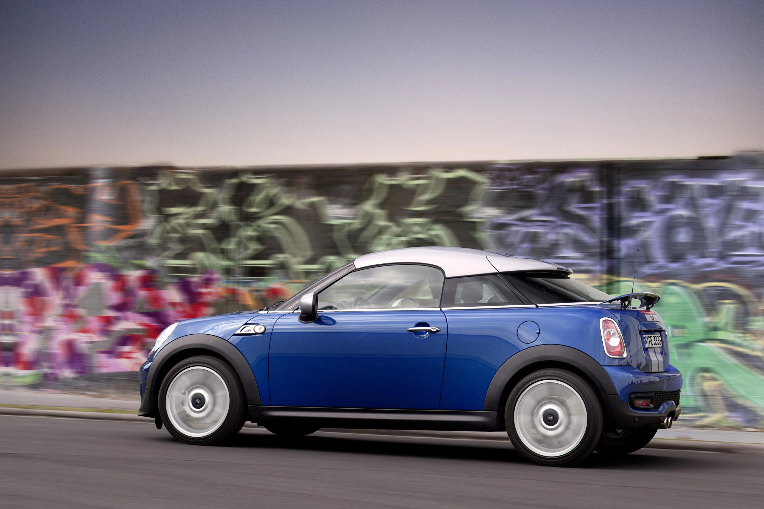Blauer Mini Cooper fährt schnell auf einer Straße, im Hintergrund eine mit Graffiti bedeckte Wand.