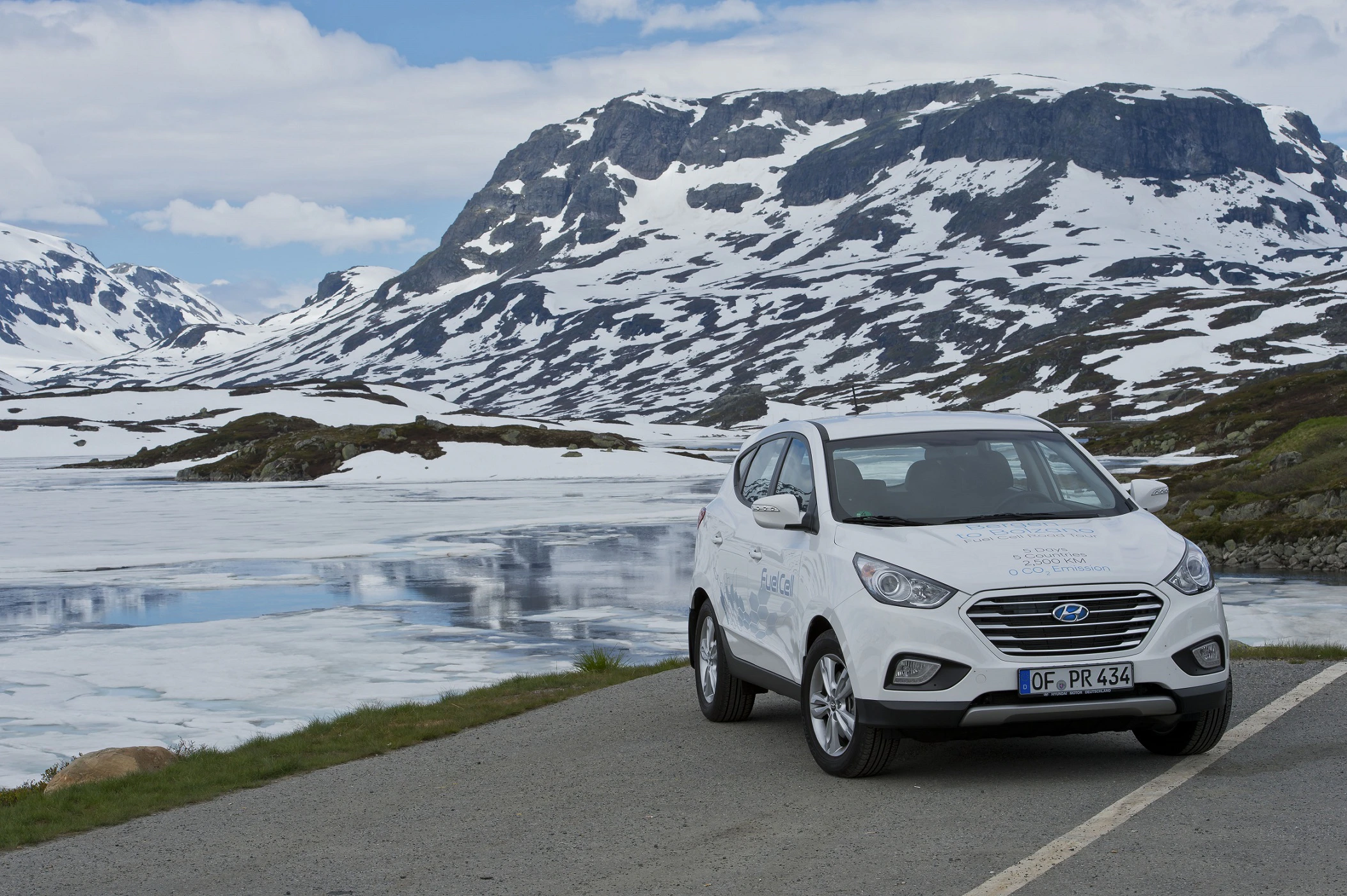 Ein weißes Auto steht auf einer Straße vor verschneiten Bergen und einem zugefrorenen See unter blauem Himmel.