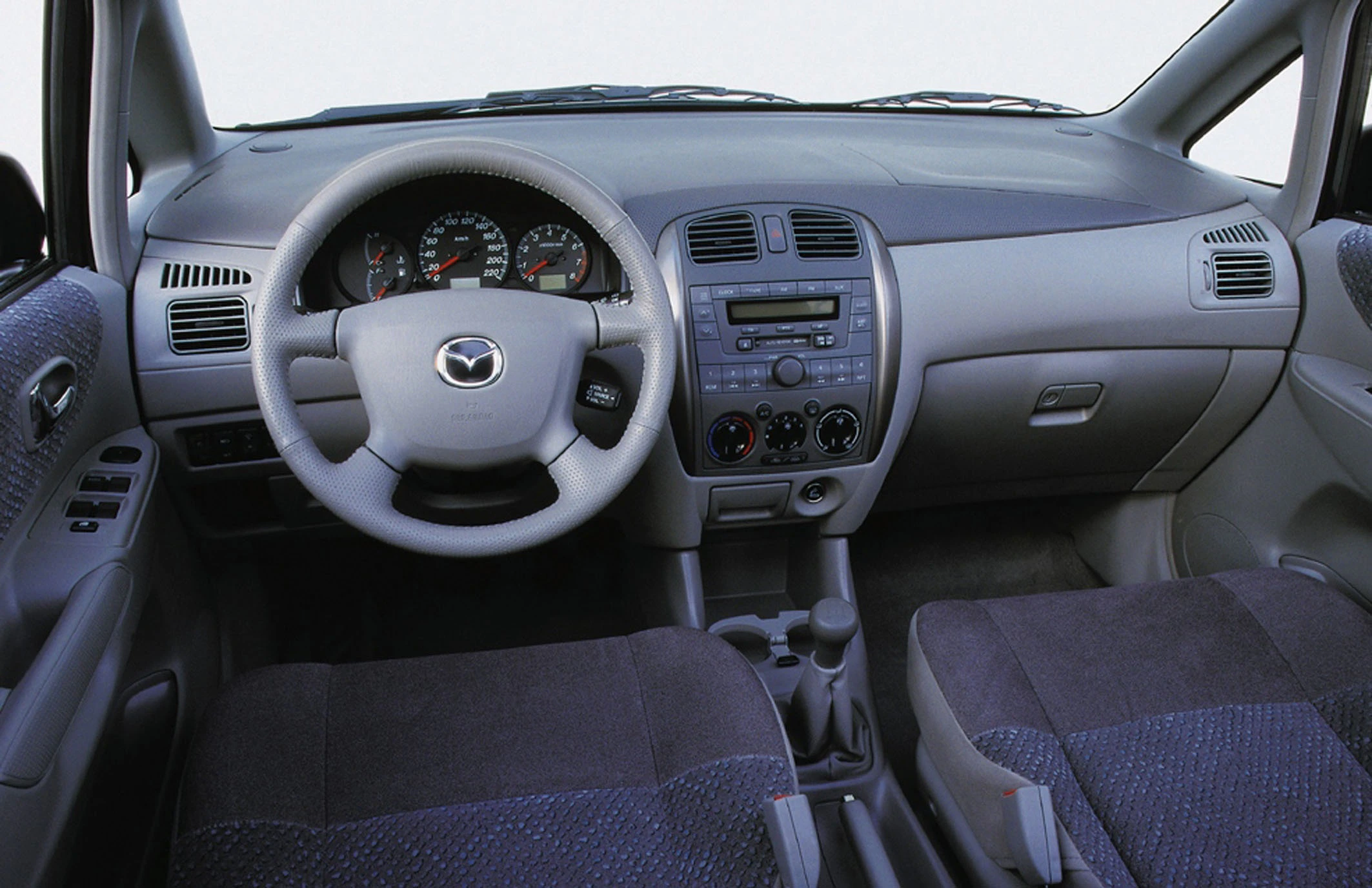 Innenraum eines Autos mit grauem Lenkrad, Tachometer, Mittelkonsole und Radiobedienfeld. Vordersitze mit grauem Stoffbezug erkennbar.