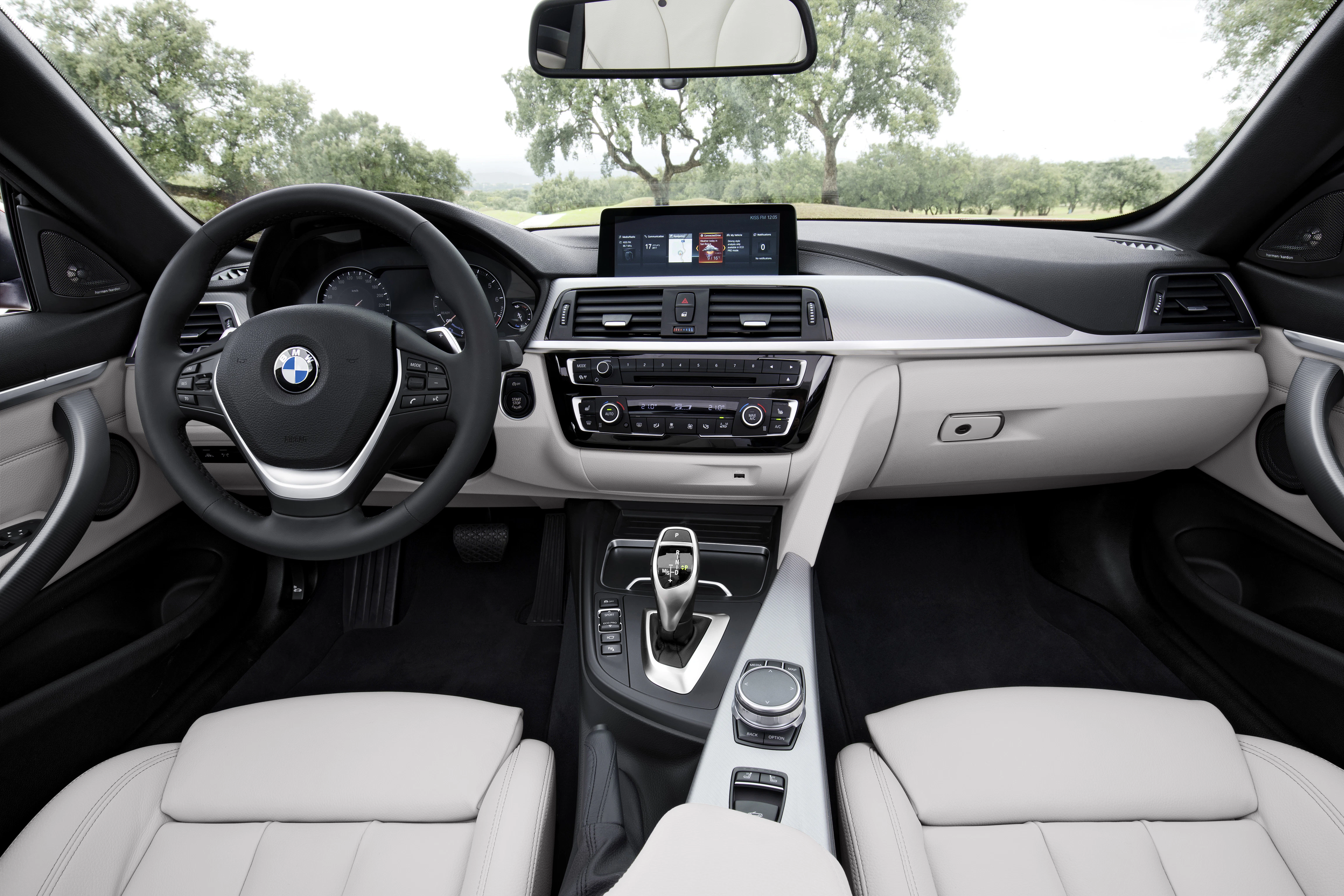 Innenraum eines modernen Autos mit Lederpolstern, multifunktionalem Lenkrad, großem Touchscreen und Schaltknopf. Außen Bäume sichtbar.