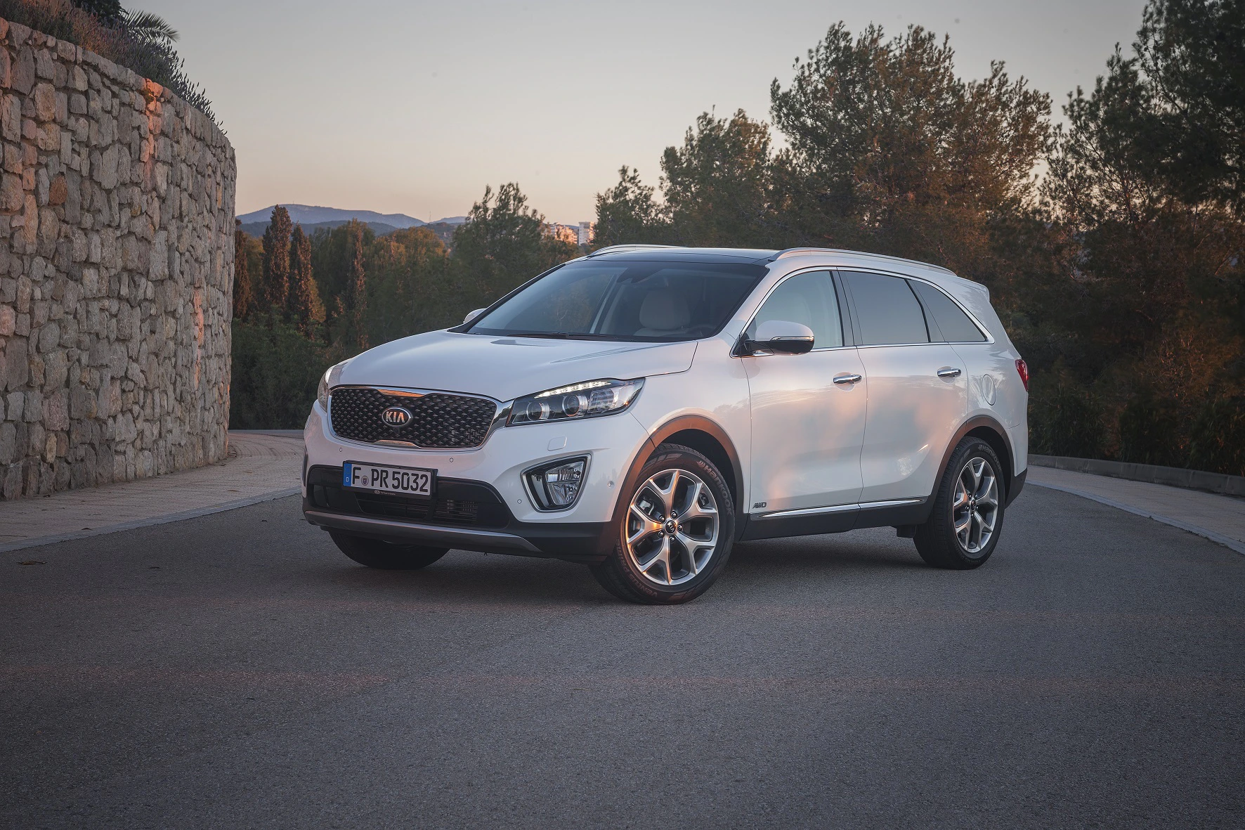 Weißer SUV parkt auf einer Straße vor einer steinernen Mauer und Bäumen, beleuchtet von sanftem Sonnenuntergangslicht.