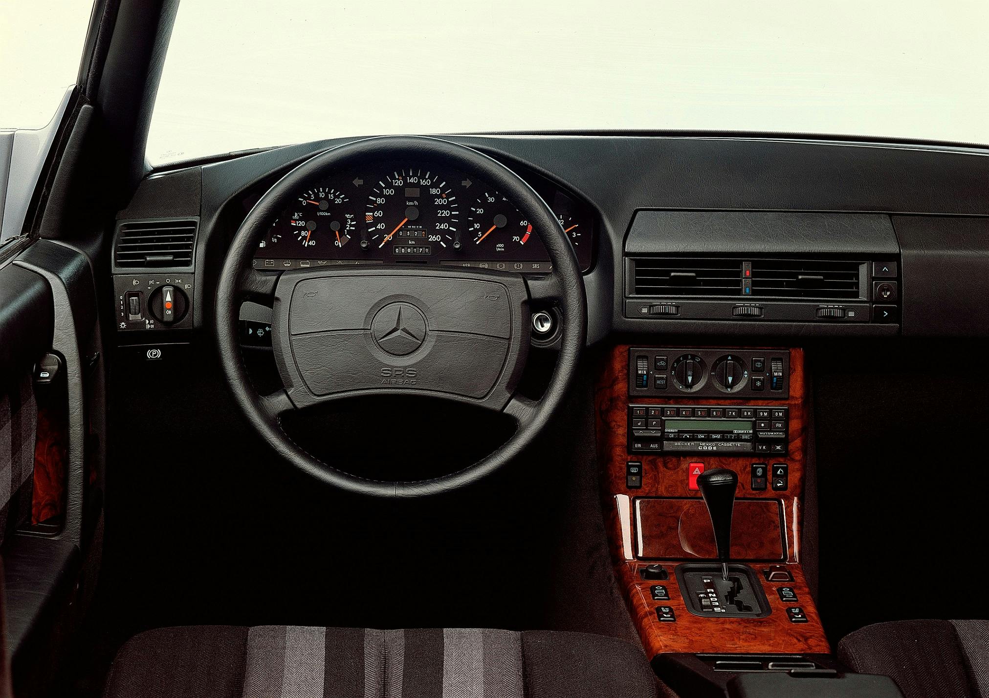 Innenraum eines klassischen Mercedes mit schwarz-grauem Armaturenbrett, Holzverkleidung, Lenkrad mit Mercedes-Logo und analogem Tachometer.