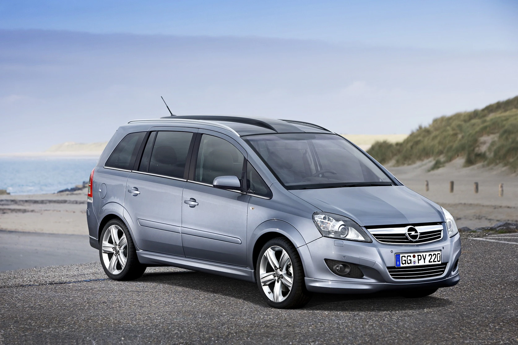 Silberner Opel Zafira geparkt an einer Küstenstraße mit Dünen im Hintergrund; sonniger Himmel.