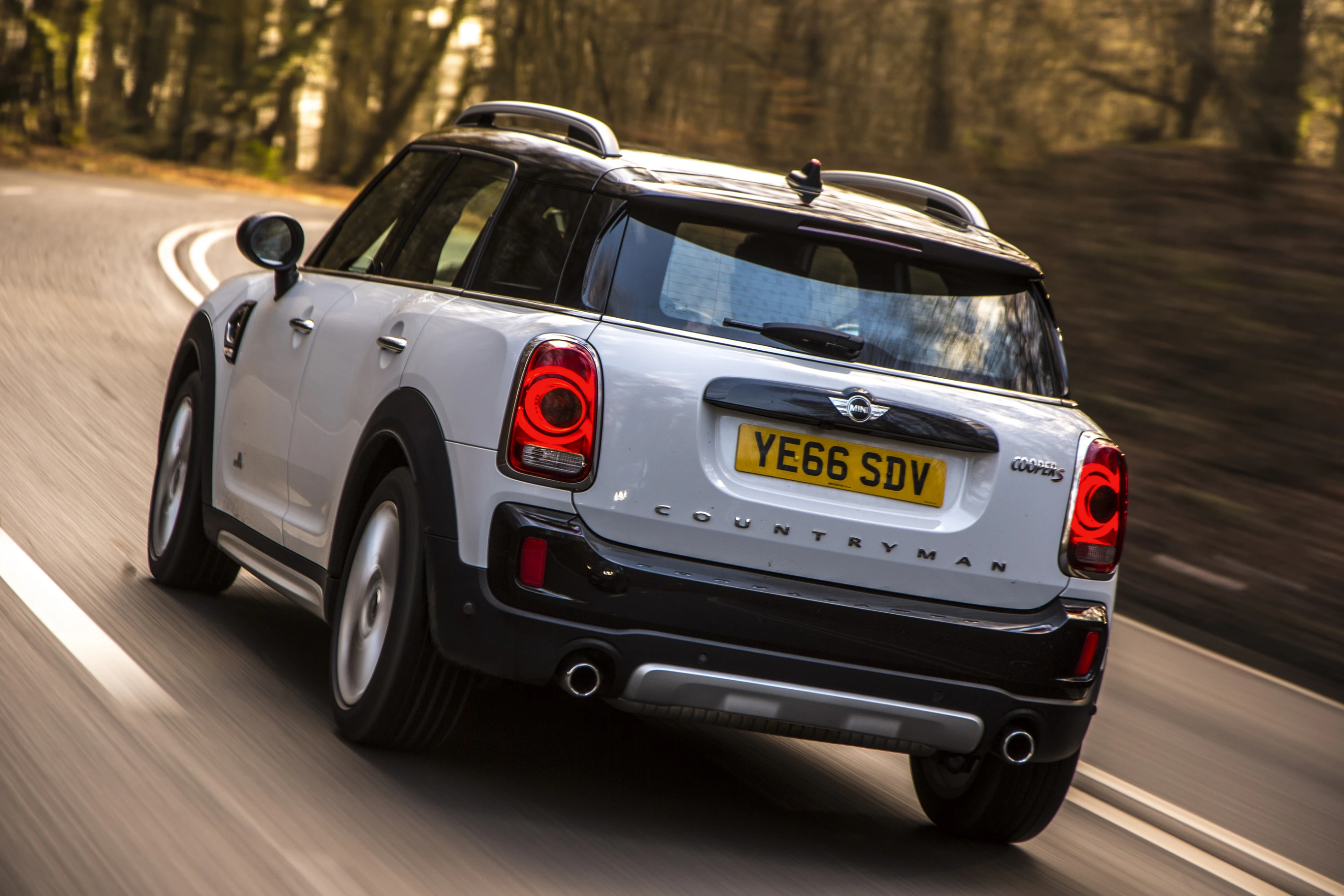 Weißer Mini Countryman fährt auf einer kurvigen Landstraße. Die Rücklichter leuchten, und um die Straße sind Bäume sichtbar.