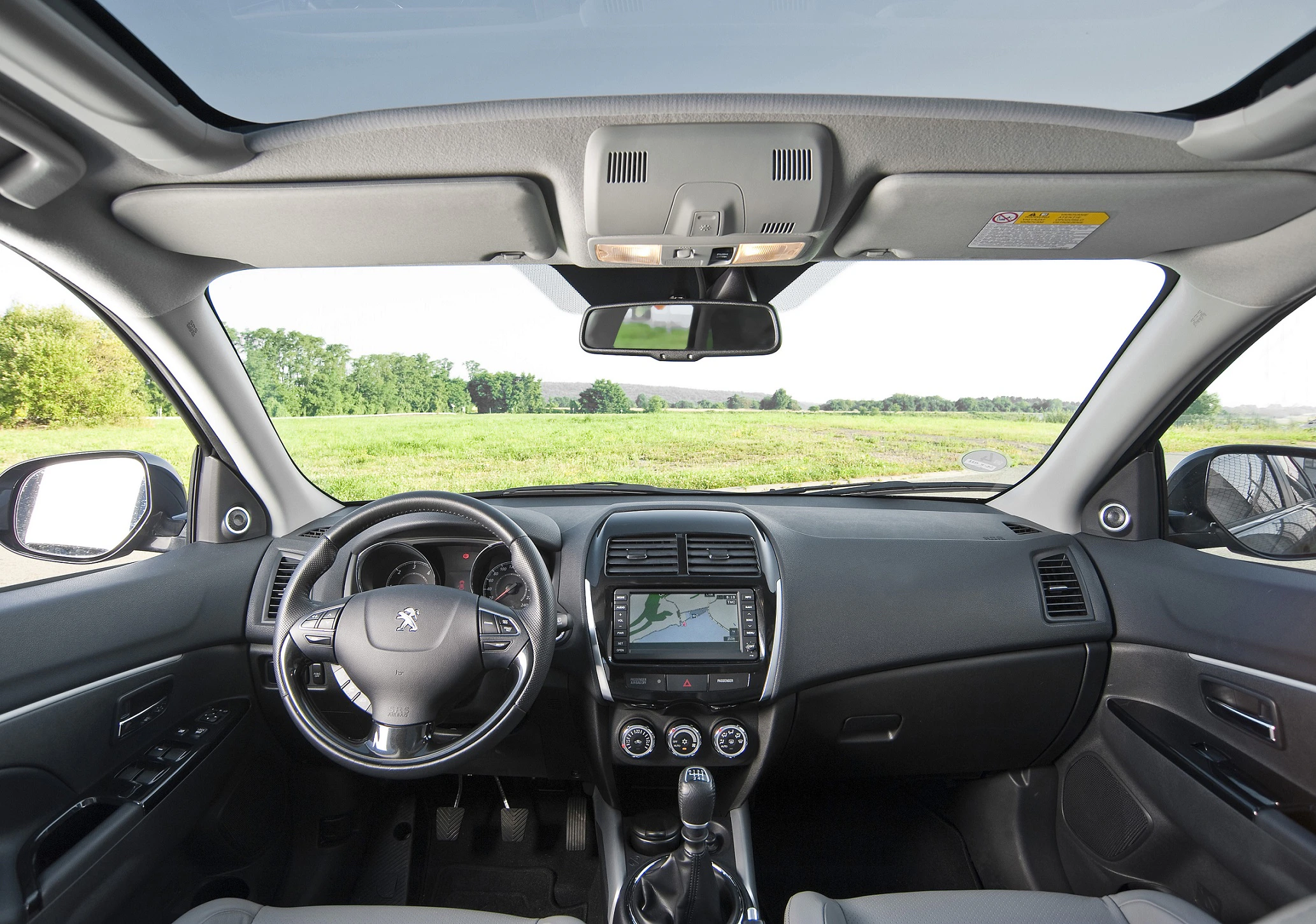 Innenraum eines Autos mit Blick durch die Frontscheibe auf ein grünes Feld. Das Lenkrad zeigt das Peugeot-Logo, ein Navigationssystem ist sichtbar.