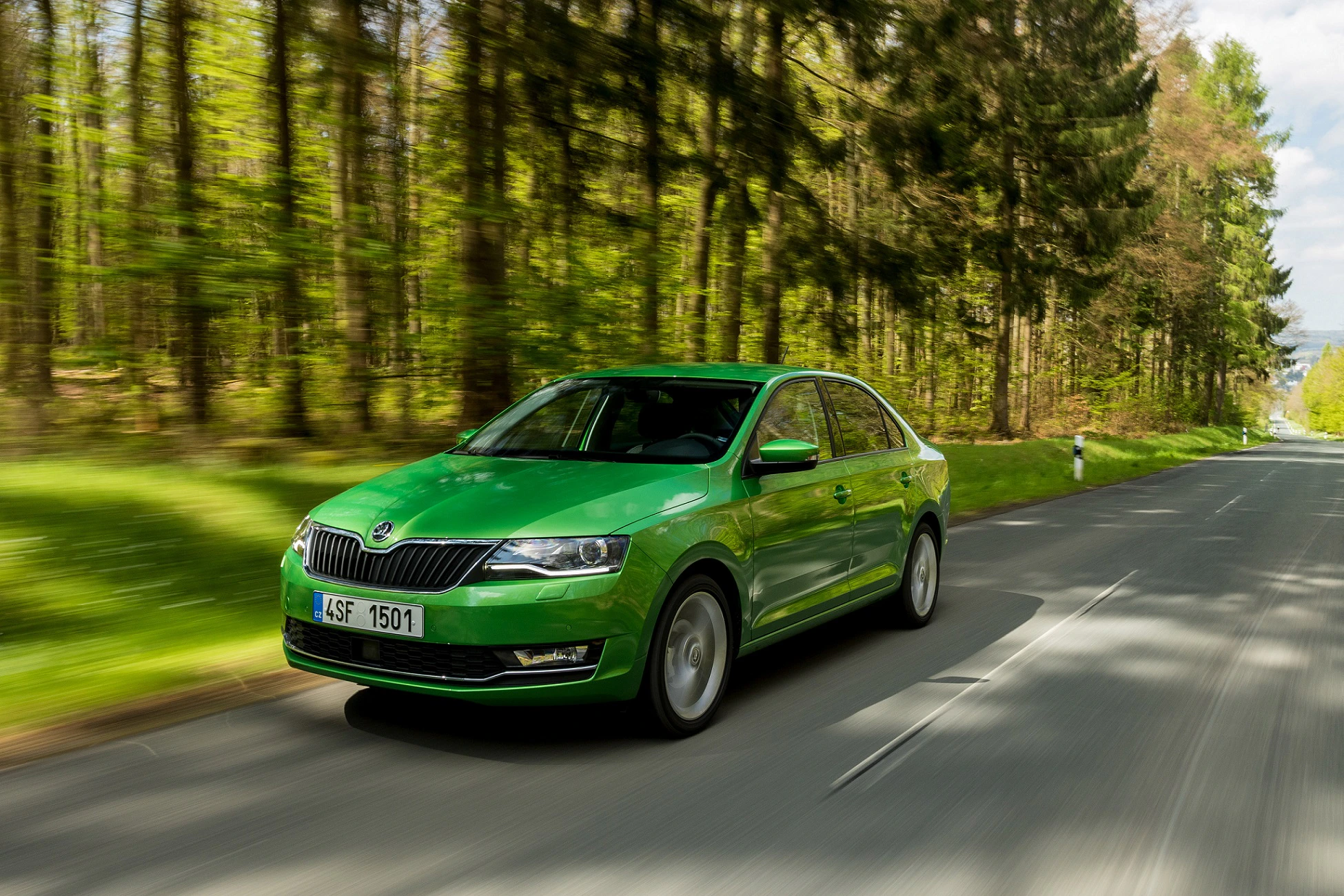 Grünes Auto fährt auf einer Landstraße durch einen Wald bei sonnigem Wetter.