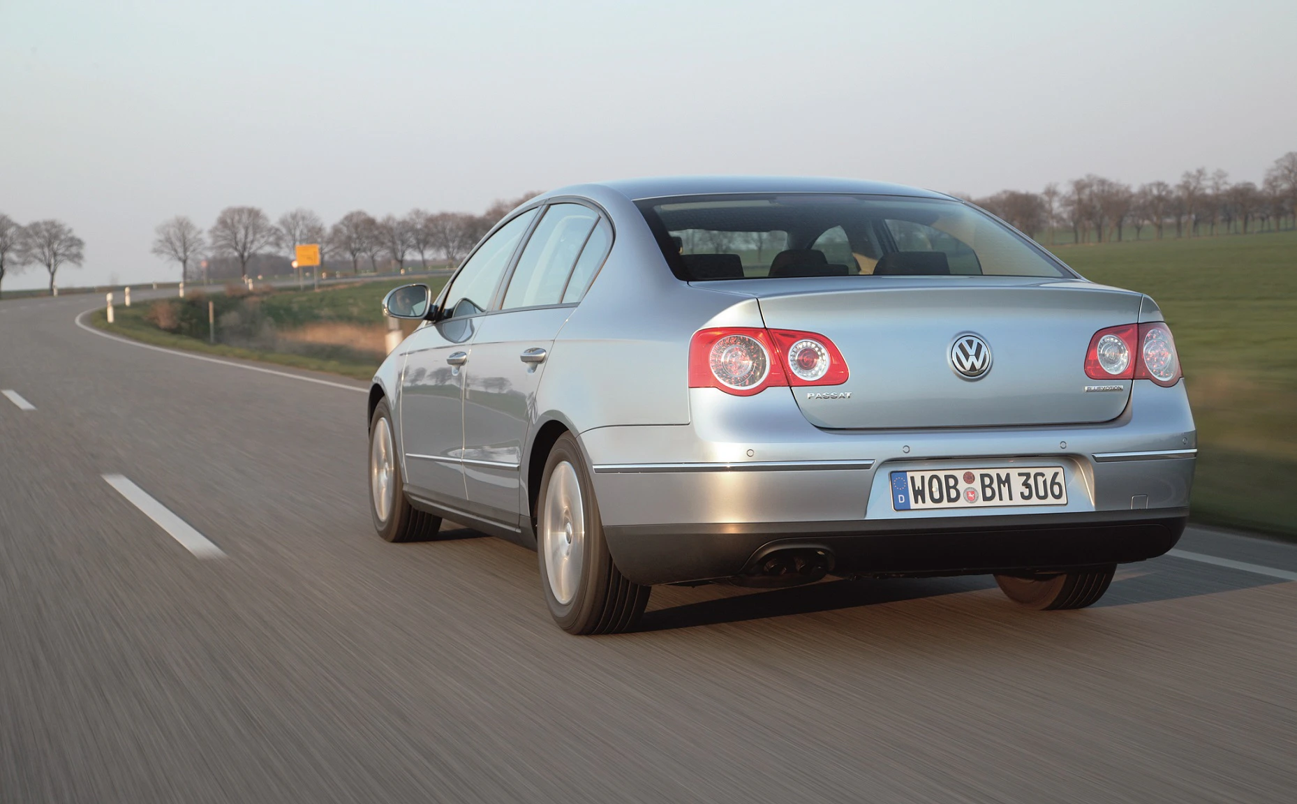 Silberner Volkswagen auf einer Landstraße, umgeben von Feldern und Bäumen bei Tageslicht. Rückansicht des Fahrzeugs.