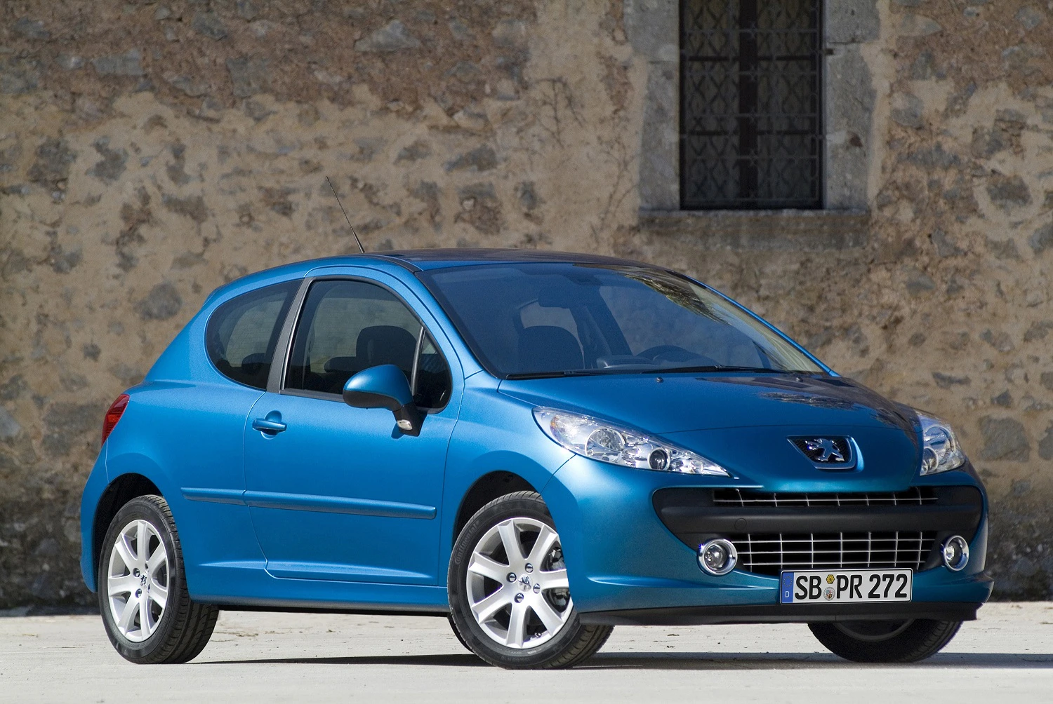 Blauer Peugeot 207 vor einer steinernen Wand geparkt, Ansicht von der Seite und vorne, mit klar sichtbarem Nummernschild.