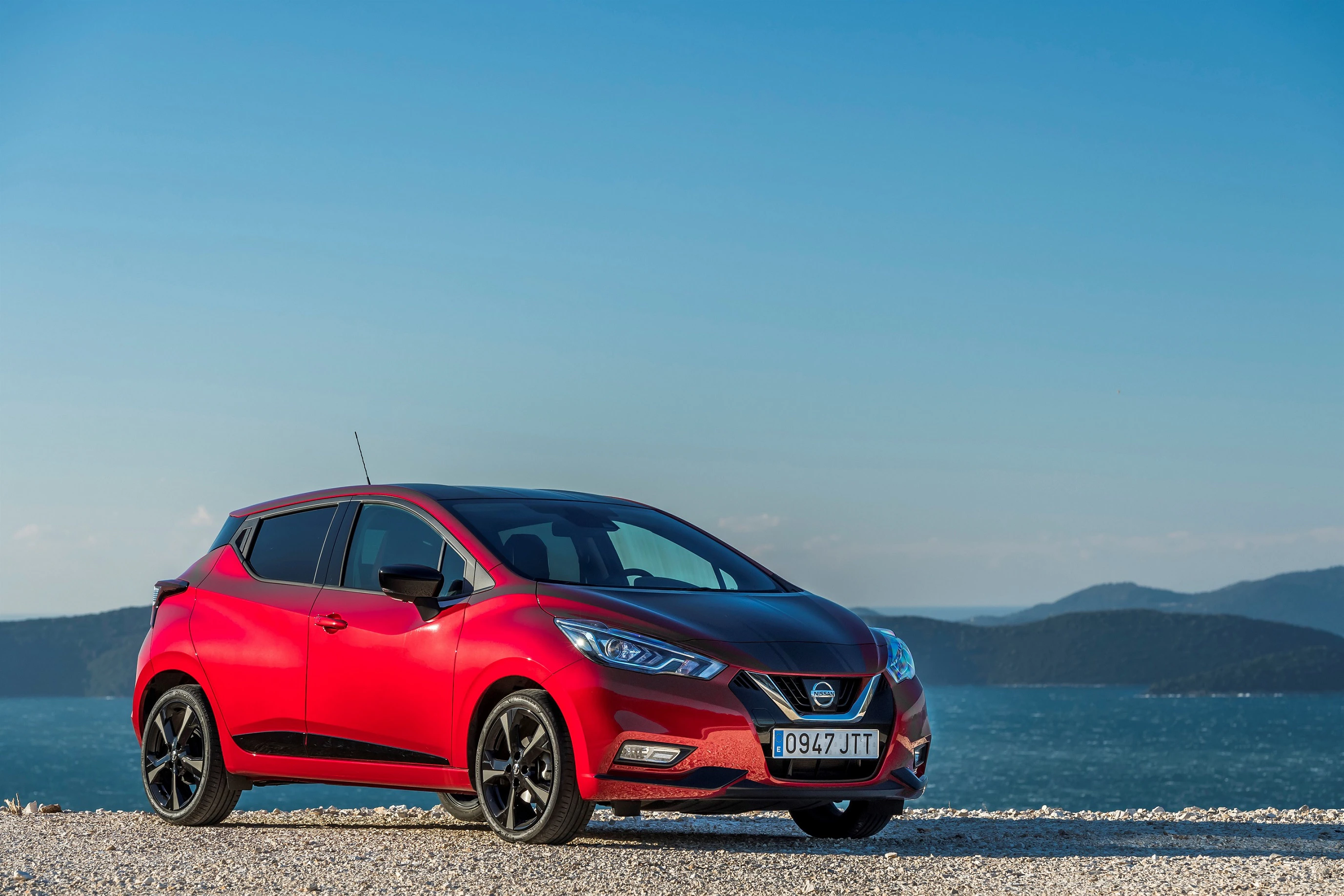 Roter Nissan Kleinwagen parkt auf einer Kiesstraße vor blauem Meer unter klarem Himmel. Im Hintergrund sind sanfte Hügel zu sehen.