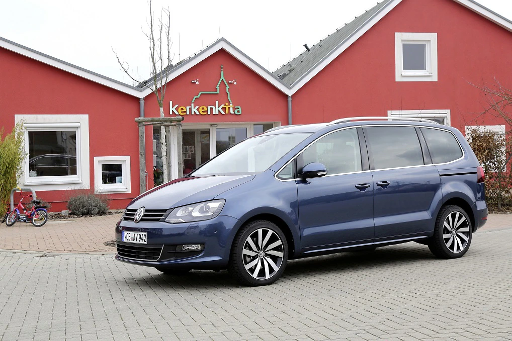 Blauer Volkswagen Sharan steht auf einem Parkplatz vor einem roten Gebäude mit dem Schriftzug "Kerkenkita". Im Hintergrund ein Dreirad.
