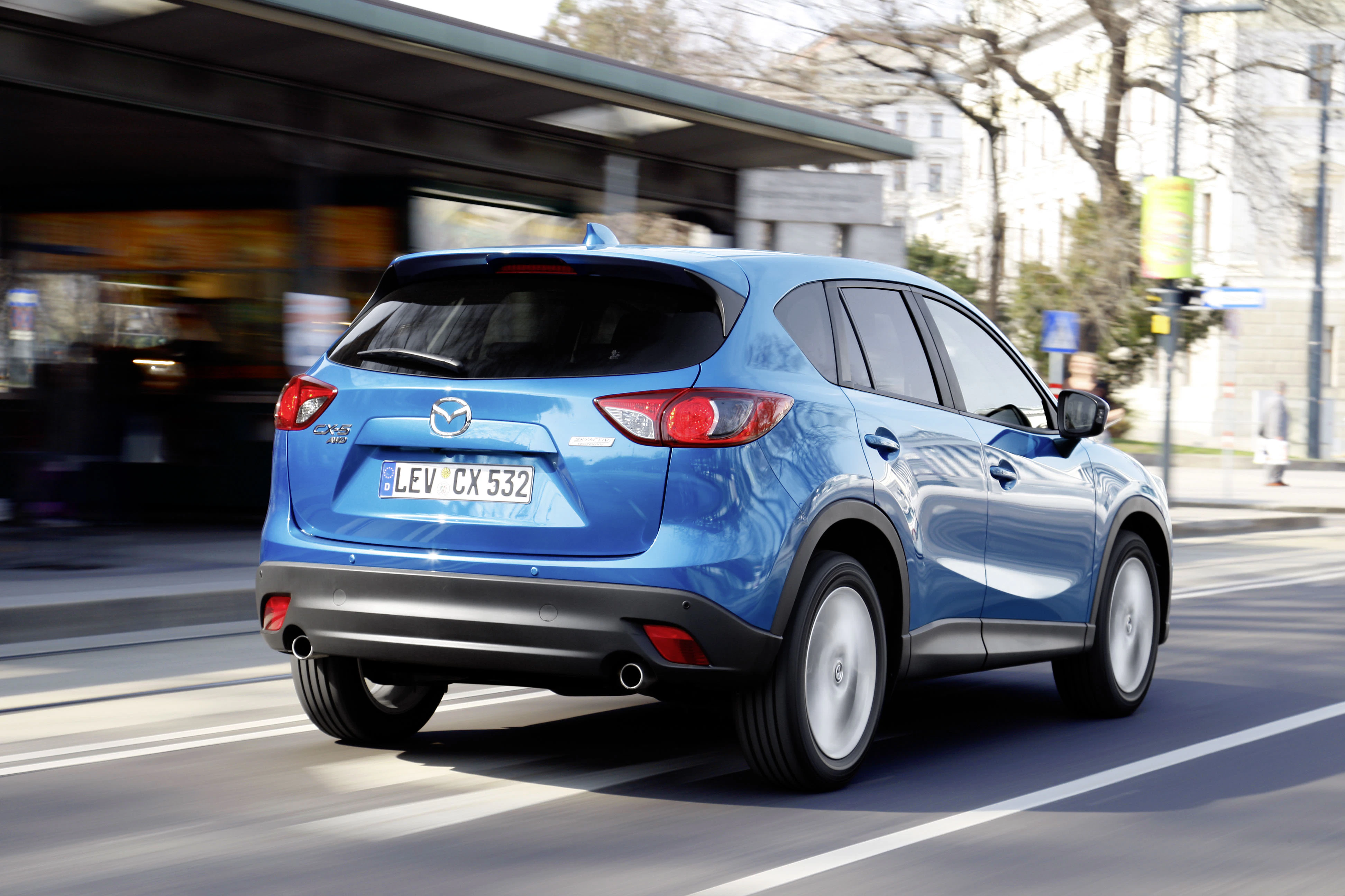 Blauer SUV von Mazda fährt auf einer städtischen Straße. Im Hintergrund sind unscharfe Gebäude und Bäume zu sehen.