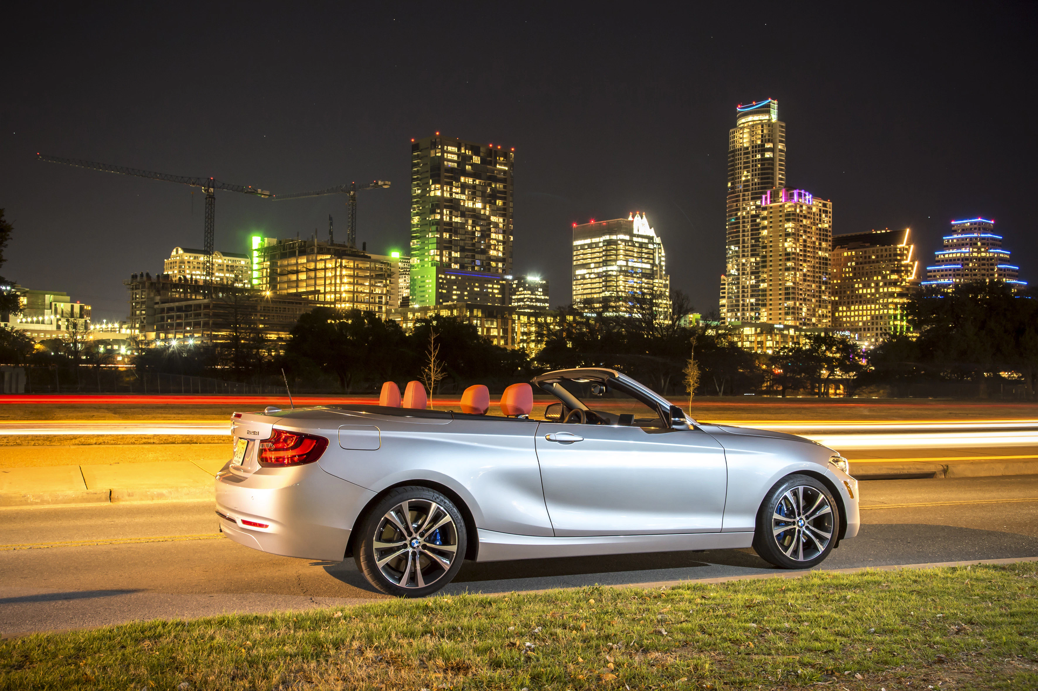 Silbernes Cabriolet parkt nachts vor erleuchteter Skyline, leuchtende Autospuren im Hintergrund.