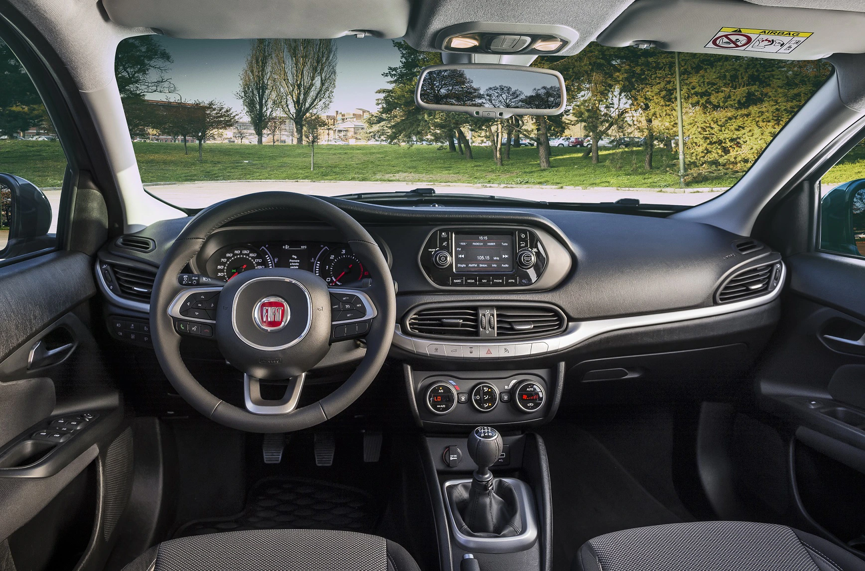 Innenraum eines Fiat-Autos mit Lenkrad, Armaturenbrett, Schalthebel und Touchscreen. Draußen grüne Bäume und Wiese sichtbar.