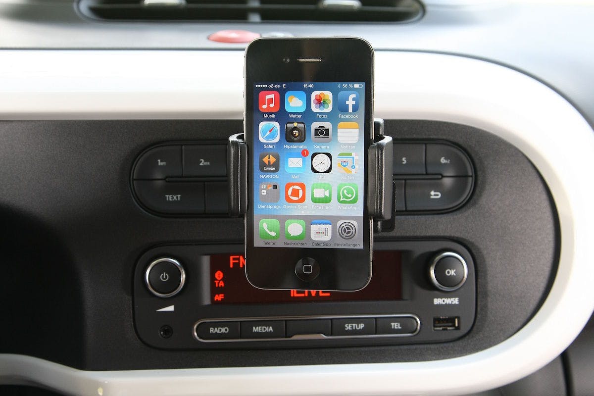 Smartphone in Halterung im Auto, zeigt Homescreen mit Apps wie Nachrichten, Safari und WhatsApp. Darunter Autoradio mit Knöpfen für Radio und Medien.