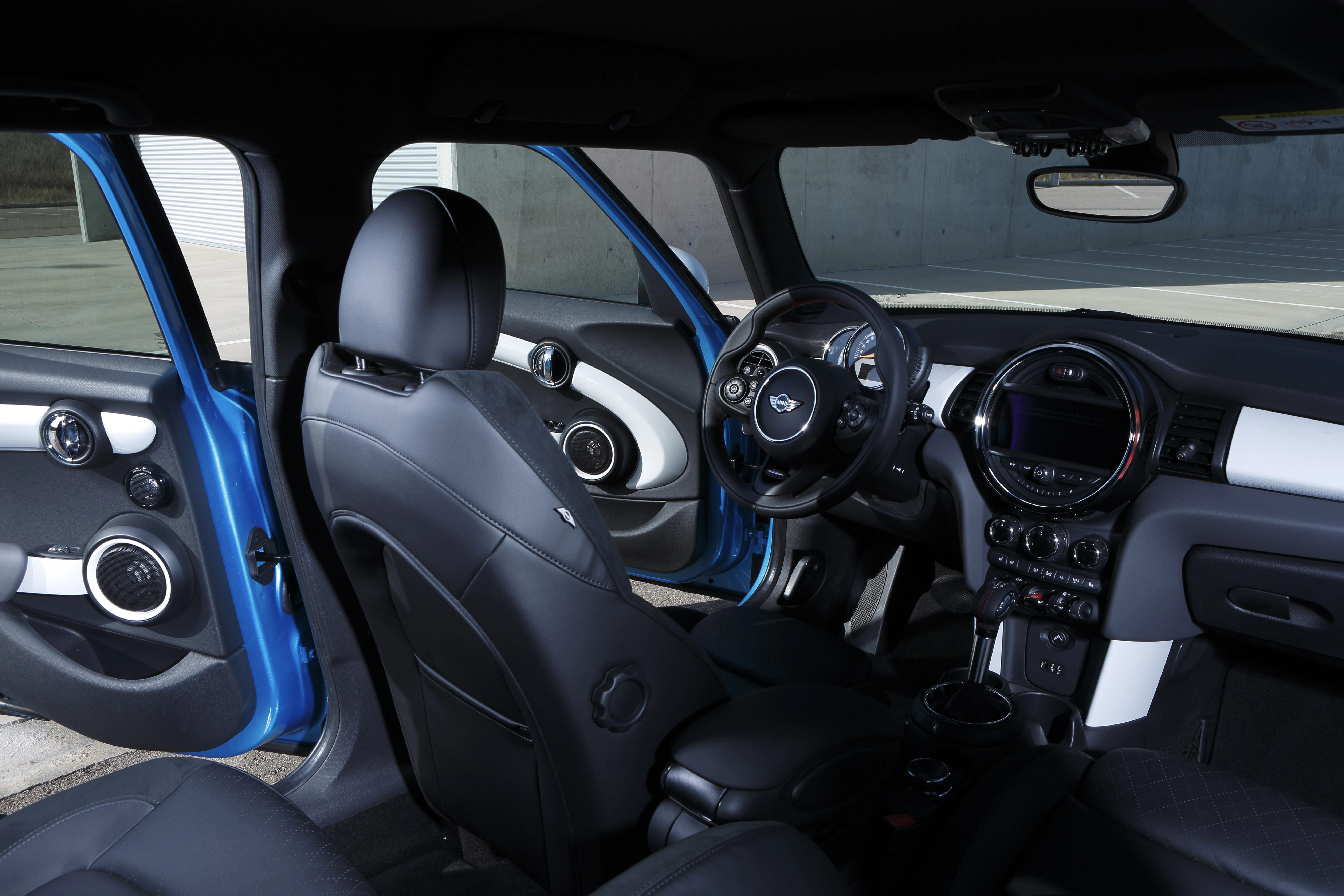 Innenraum eines Autos mit modernem Lenkrad, blauen Türen und schwarzen Ledersitzen, aufgenommen bei geöffneter Fahrertür.