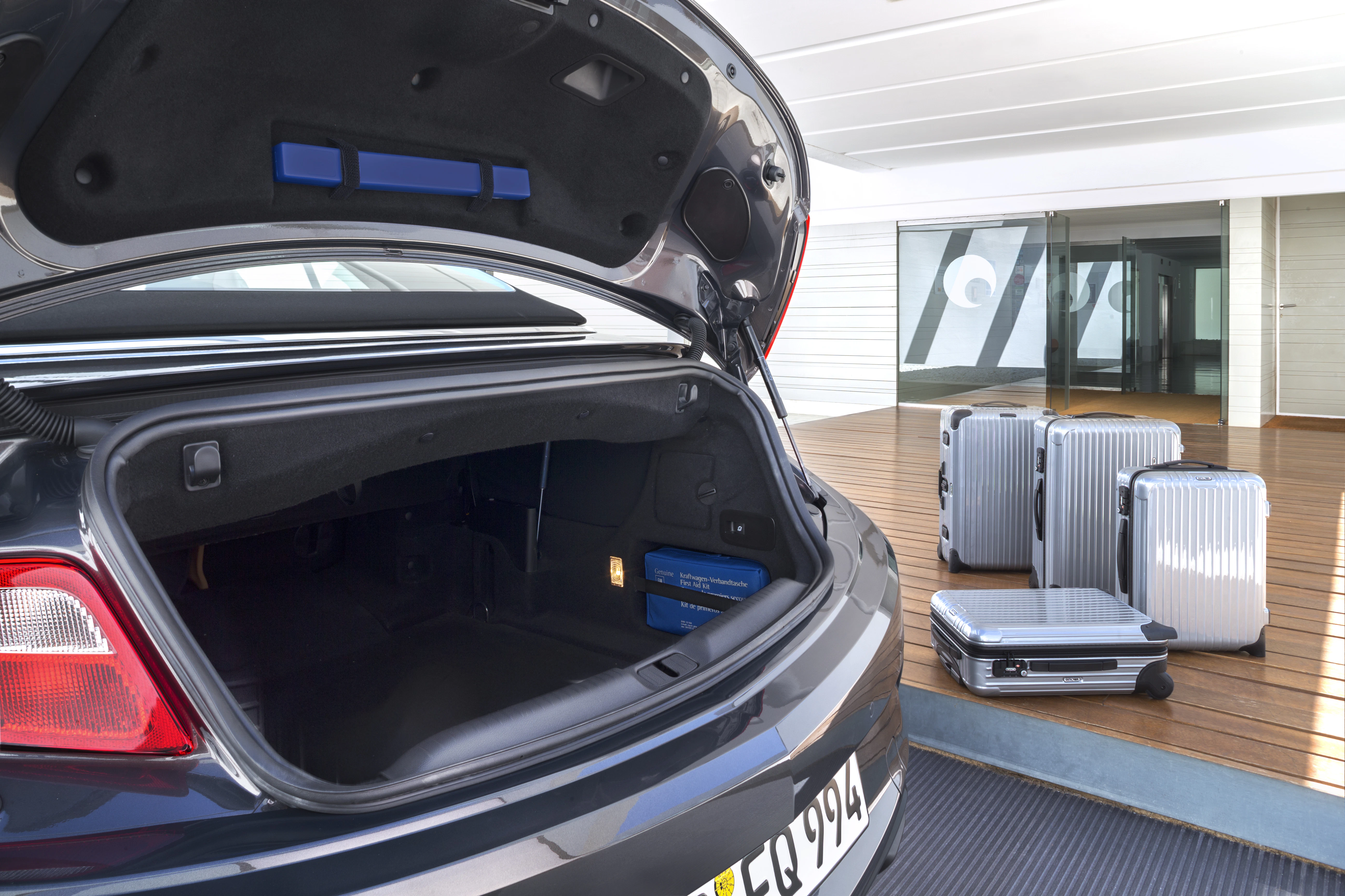 Offener Kofferraum eines Autos, daneben drei silberne Koffer auf einem Holzdeck vor einem modernen Gebäude.