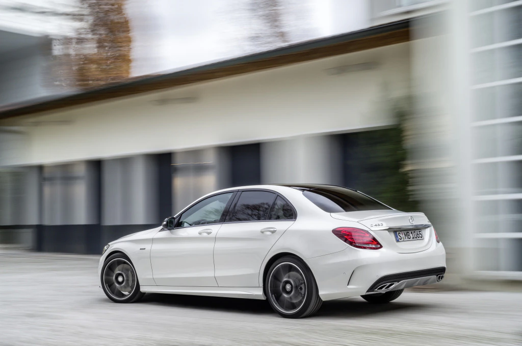 Weißer Mercedes-Benz C-Klasse fährt dynamisch auf einer Straße entlang eines Gebäudes. Schnelligkeit und Eleganz werden betont.