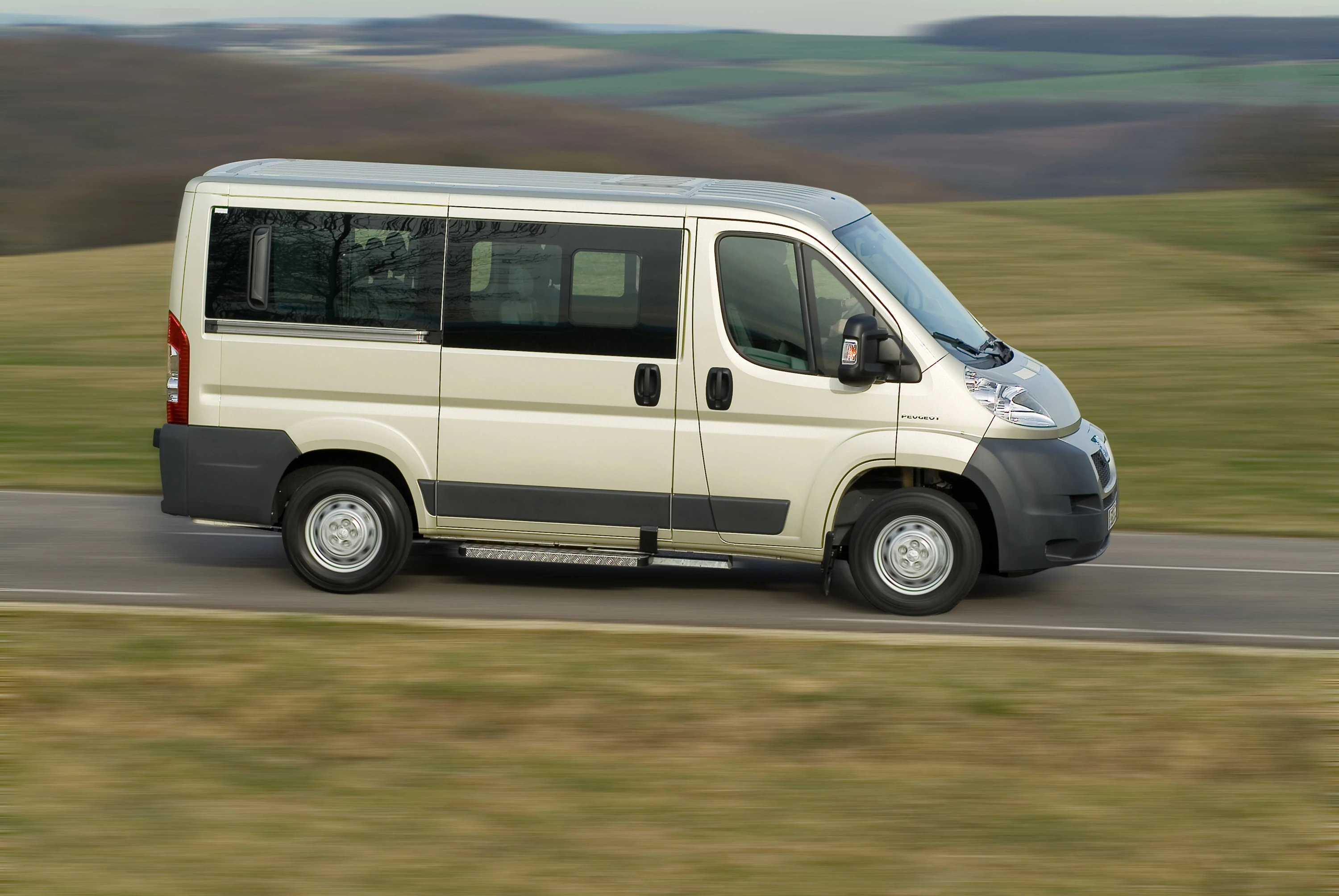 Ein weißer Kleinbus fährt schnell auf einer Landstraße durch eine ländliche, hügelige Landschaft.