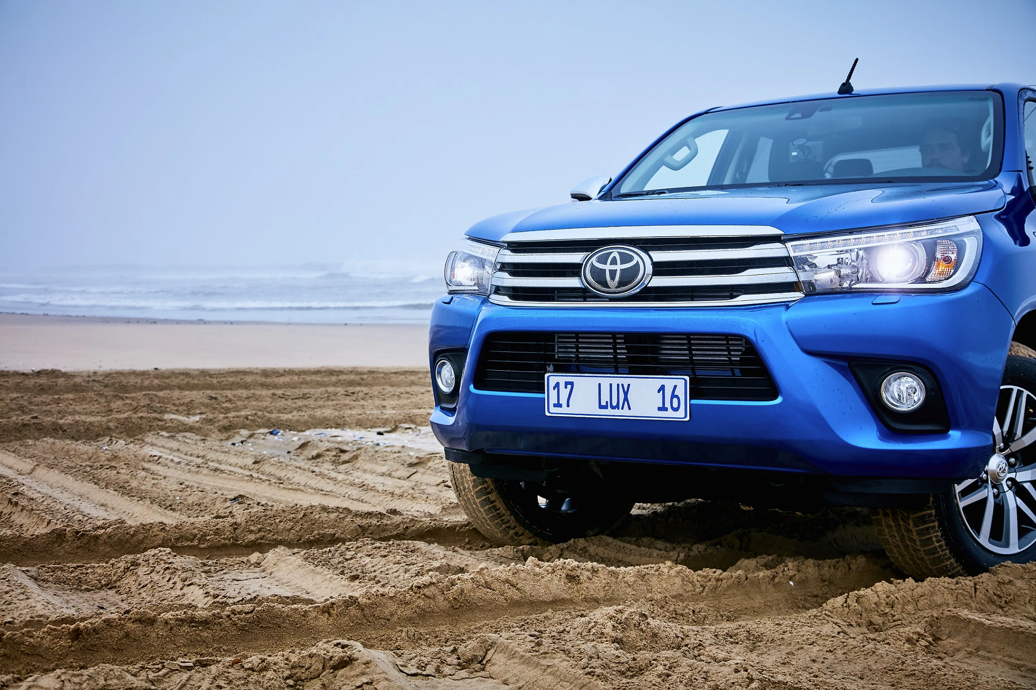 Blauer Toyota-Geländewagen fährt auf sandigem Strand, Meer im Hintergrund, bewölkter Himmel.