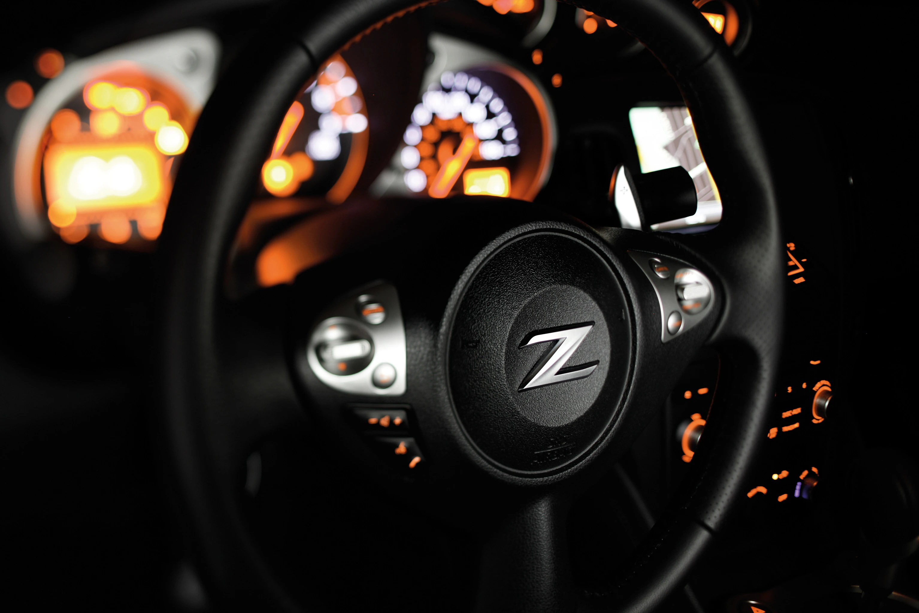 Nahaufnahme eines Autolenkrads mit Z-Logo, umgeben von orange beleuchteten Armaturenbrettanzeigen.