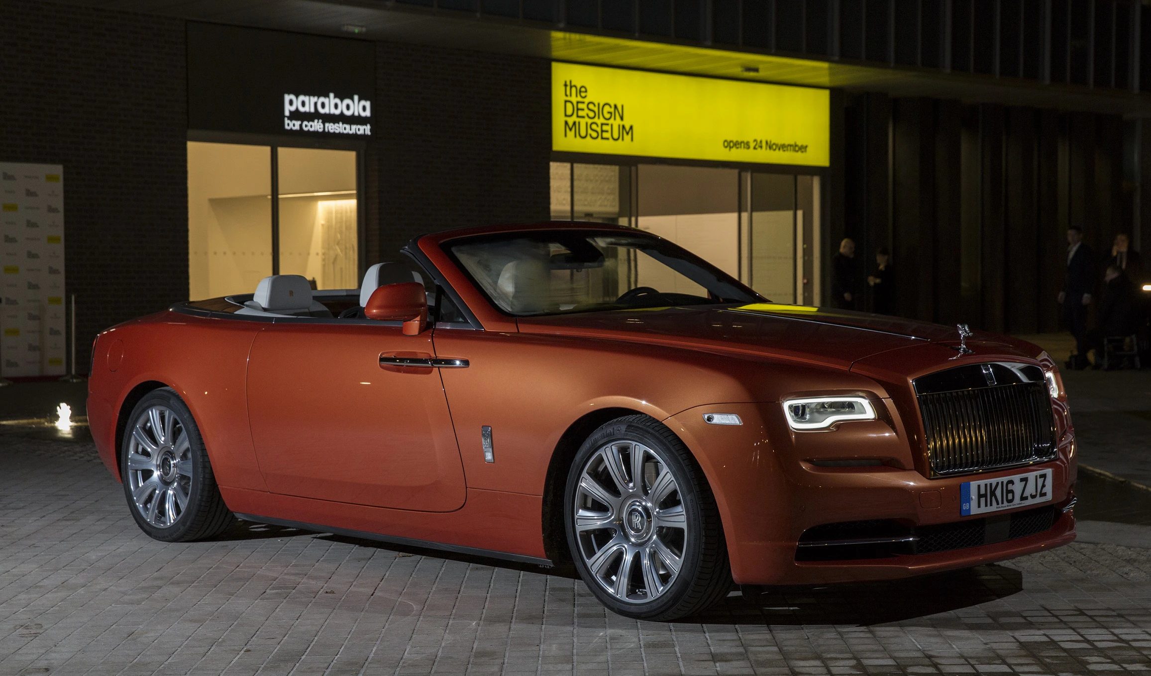 Ein elegantes, orangefarbenes Cabrio (Rolls-Royce) steht nachts vor einem beleuchteten Gebäude, das "Design Museum" anzeigt.