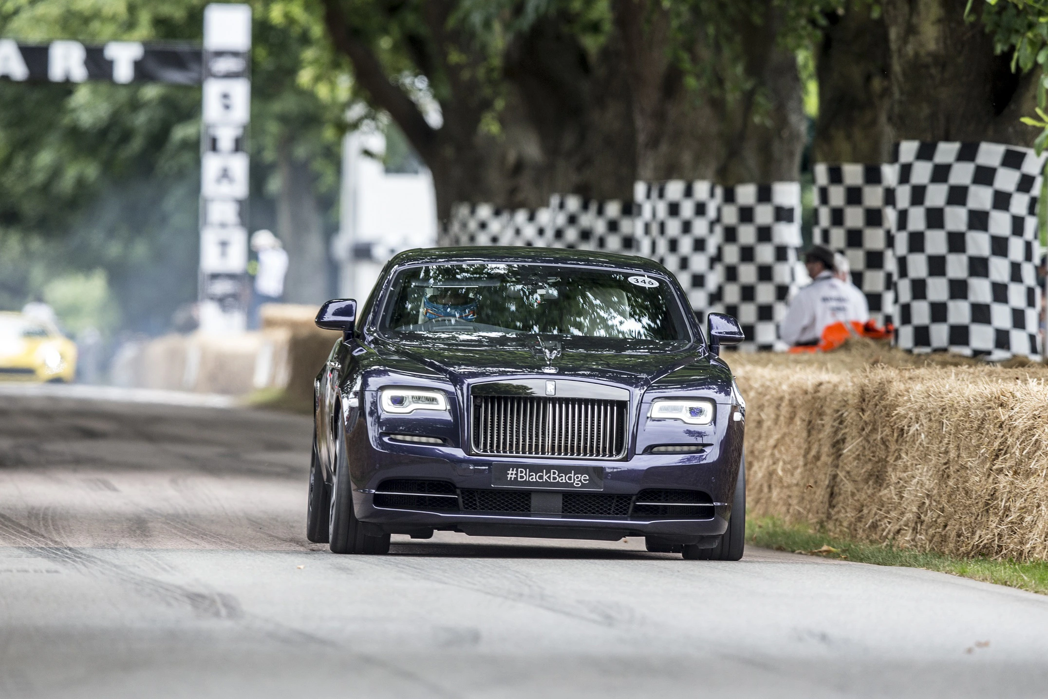Ein schwarzer Rolls-Royce fährt auf einer Straße einer Motorsport-Veranstaltung. Links und rechts sind schwarz-weiße Karomuster zu sehen.