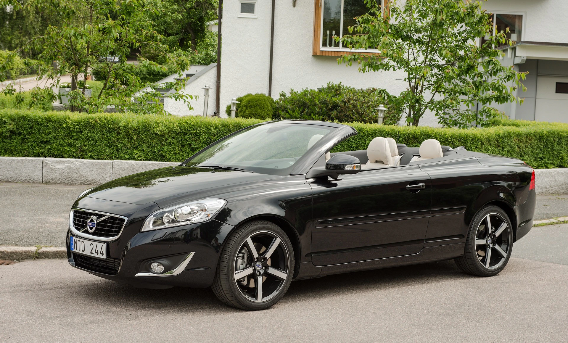 Schwarzes Volvo Cabriolet, geparkt am Straßenrand vor einem Wohnhaus mit grünem Garten im Hintergrund.