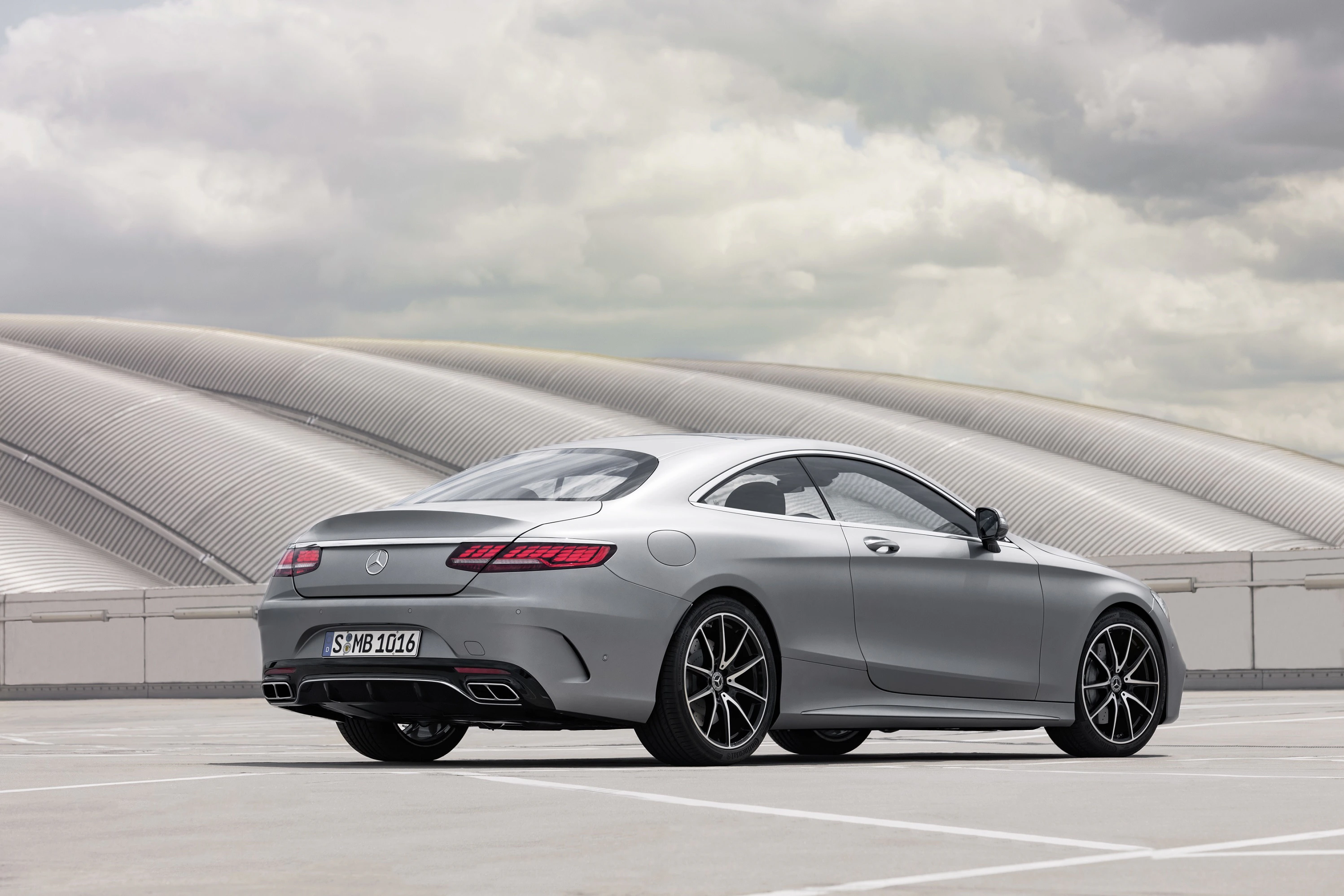 Silbernes Mercedes Coupé auf einem Parkdeck vor einer modernen, wellenförmigen Architektur unter einem bewölkten Himmel.