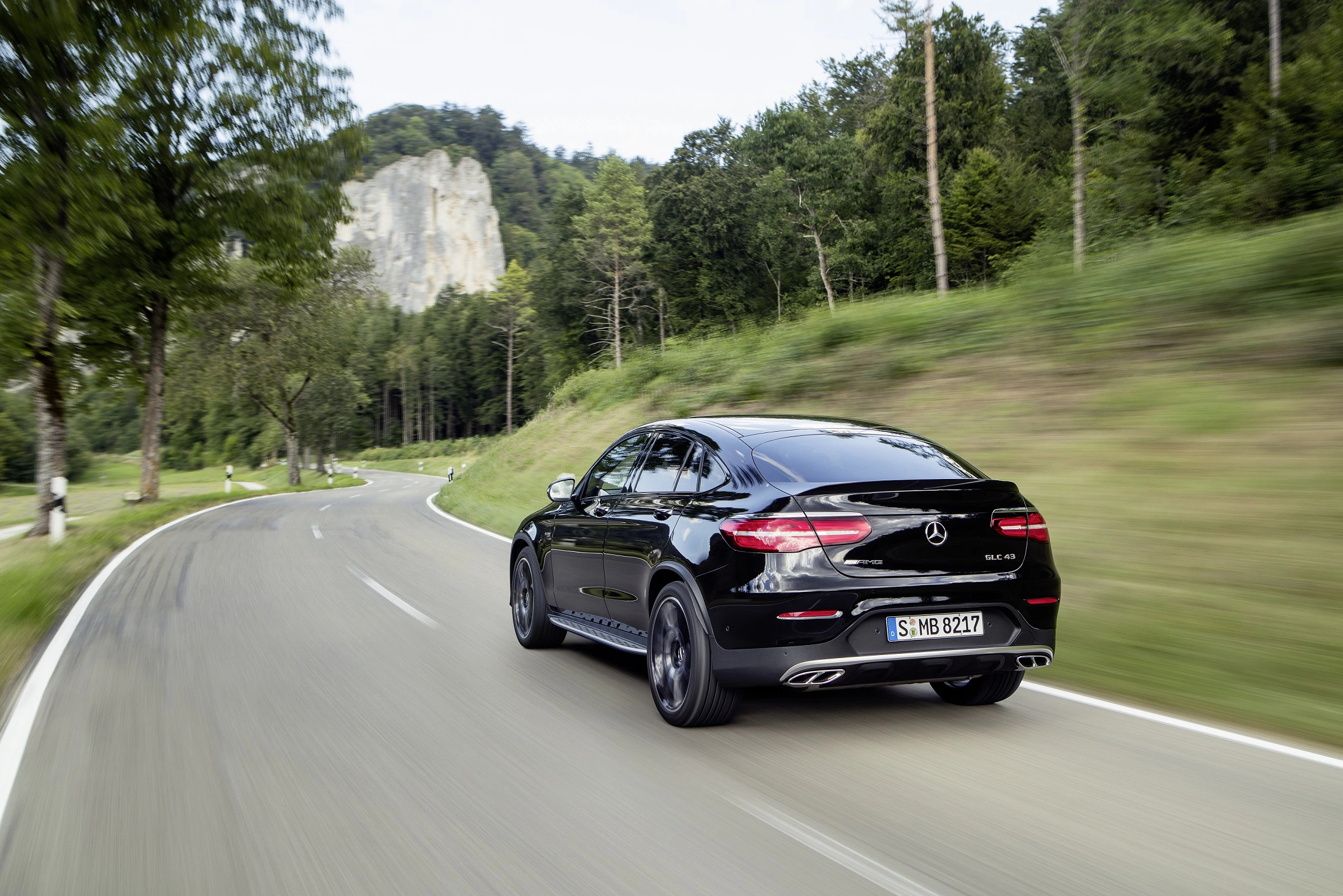 Schwarzer Mercedes-Benz fährt auf kurviger Landstraße durch waldreiche Landschaft, im Hintergrund Hügel und Felsen.