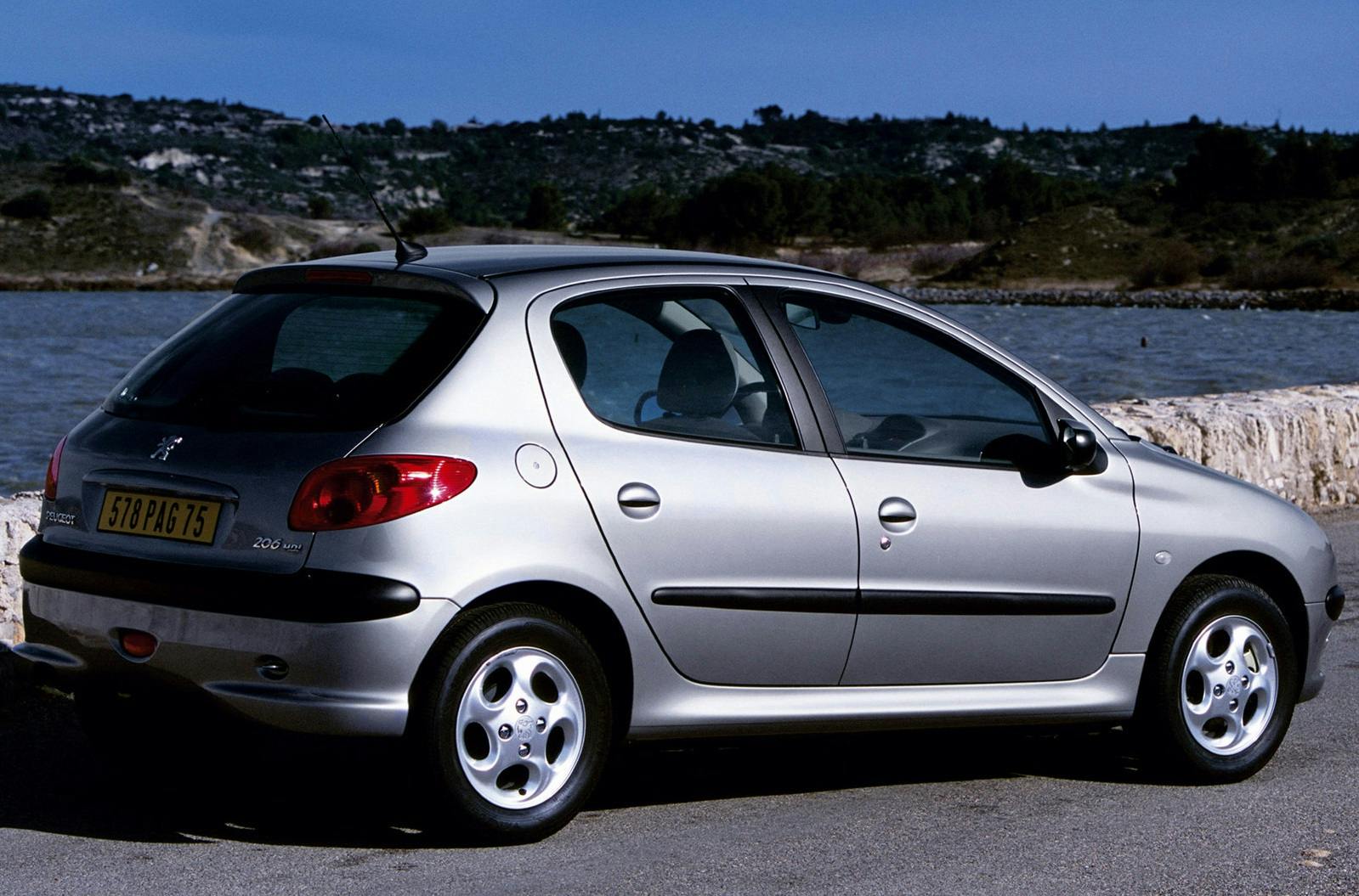 Silberner Peugeot 206 steht an einem See mit Hügeln im Hintergrund. Klarer Himmel und sonniger Tag.