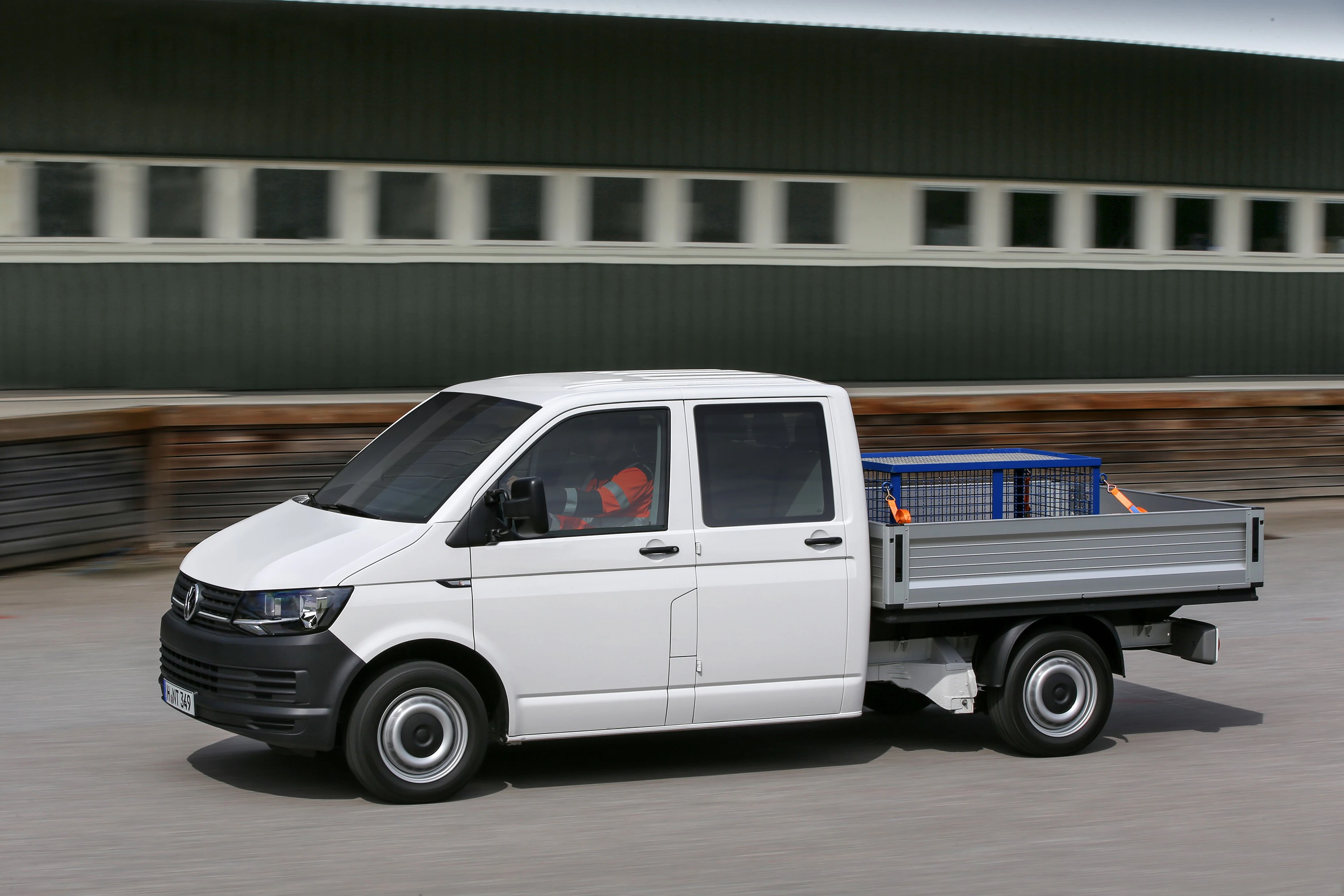 Weißer Transporter mit Pritschenaufbau fährt vor einem grünen Gebäude. Fahrer in orangener Warnweste sichtbar.